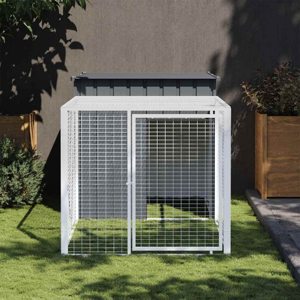 Ein weißer Drahtkäfig mit offener Tür steht auf Gras. Er hat ein graues Dach. Pflanzen und eine dunkle Wand sind im Hintergrund.
