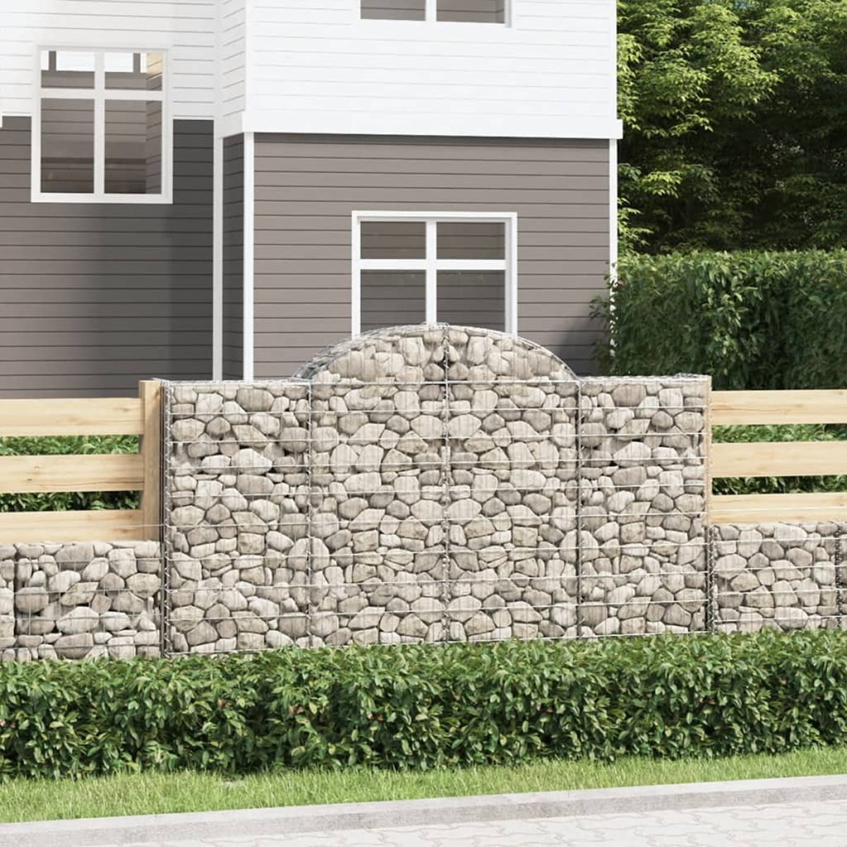 Eine Steinmauer mit Holzzaun und ein Haus mit Fenstern im Hintergrund. Grüne Büsche und Gras im Vordergrund.