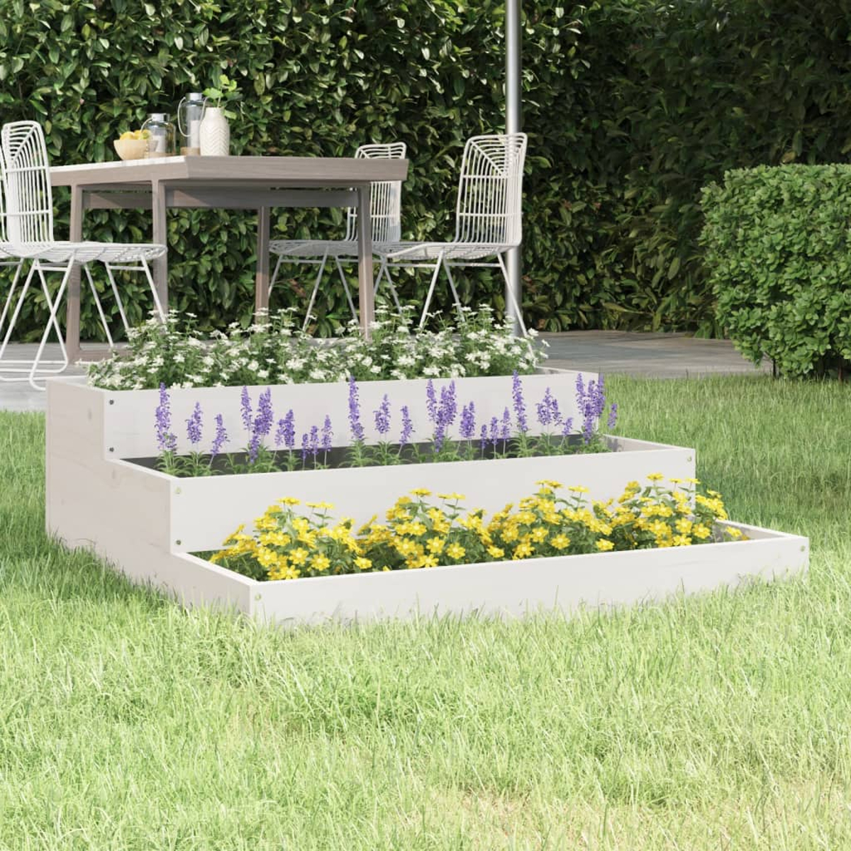 Ein gestuftes Gartenbeet mit weißen, lila und gelben Blumen steht auf einem grünen Rasen. Im Hintergrund stehen Tisch und Stühle.