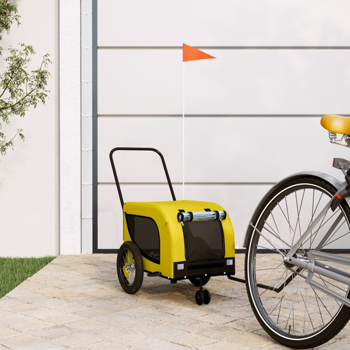 Ein gelber Fahrradanhänger für Haustiere mit einer orangefarbenen Flagge ist an einem Fahrrad befestigt. Das Fahrrad steht vor einer weißen Garage auf einem gepflasterten Bereich.