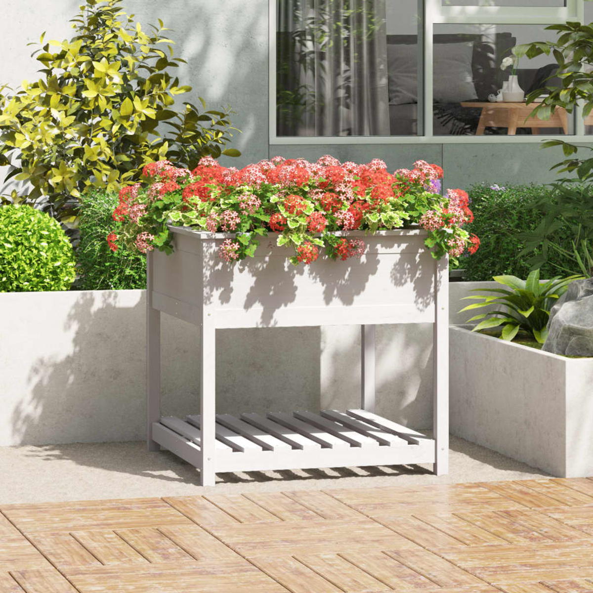 Weißer Pflanzkasten mit roten Blumen. Grüne Pflanzen umgeben den Pflanzkasten. Holzterrasse im Vordergrund und ein Gebäude im Hintergrund.