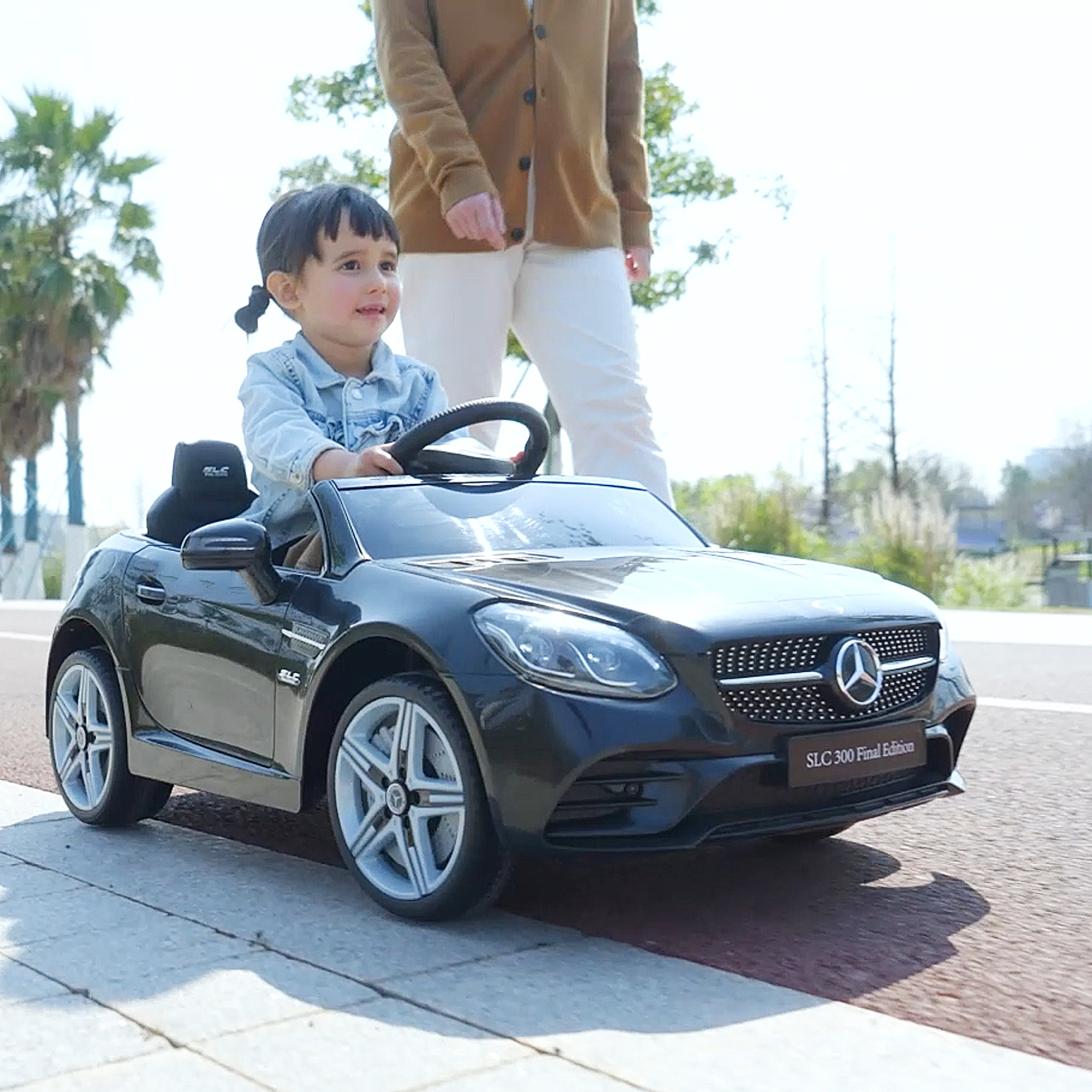 Un bambino guida un'auto giocattolo nera. Un adulto è dietro. L'auto è un modello Mercedes-Benz.