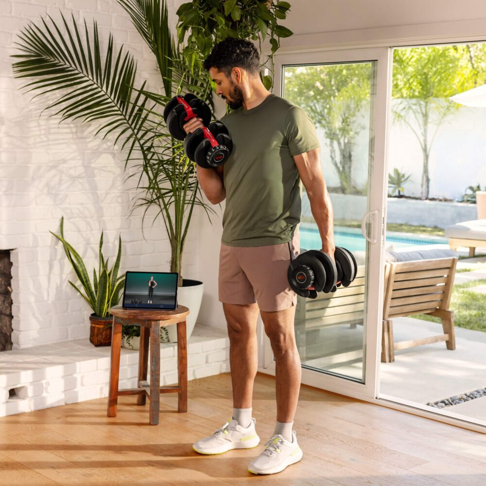 Ein Mann in grünem Hemd und Shorts hebt Gewichte. Ein Tablet zeigt ein Training. Pflanzen und ein Fenster sind im Hintergrund.
