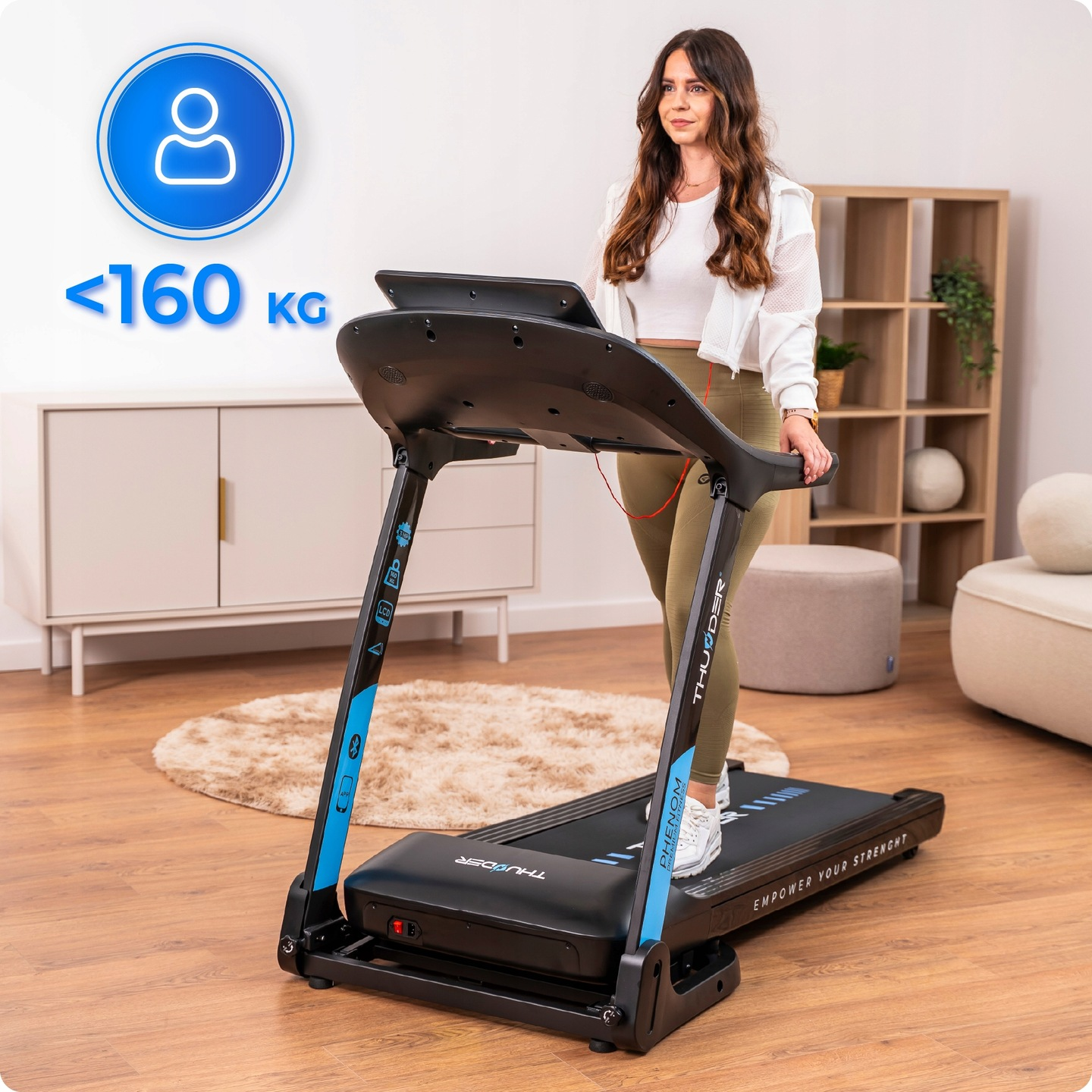 Kobieta na bieżni. Ma na sobie biały top, kurtkę i oliwkowe legginsy. Bieżnia jest czarna i niebieska. Tło obejmuje meble i dywan.