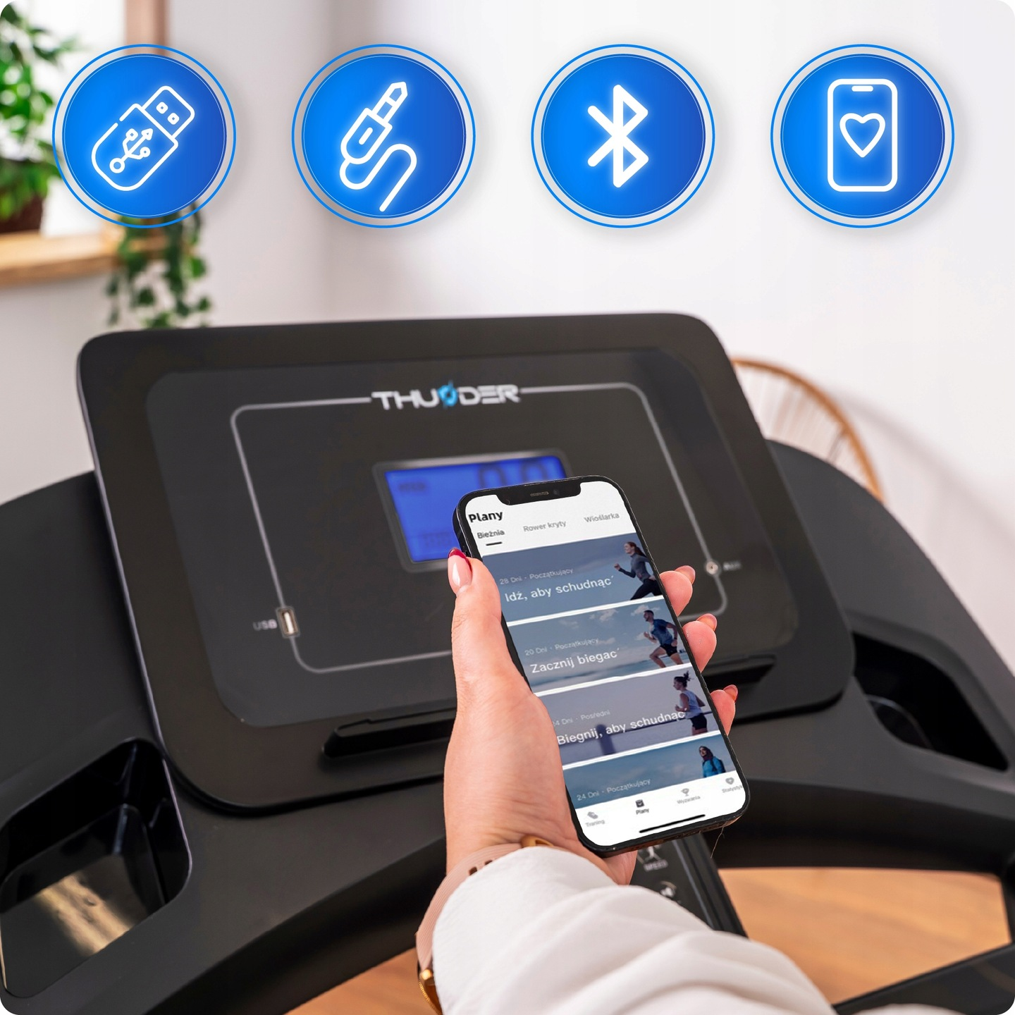 Kobieta trzyma telefon z planami treningowymi w pobliżu bieżni. Niebieskie ikony USB, jack, bluetooth i serca w tle.