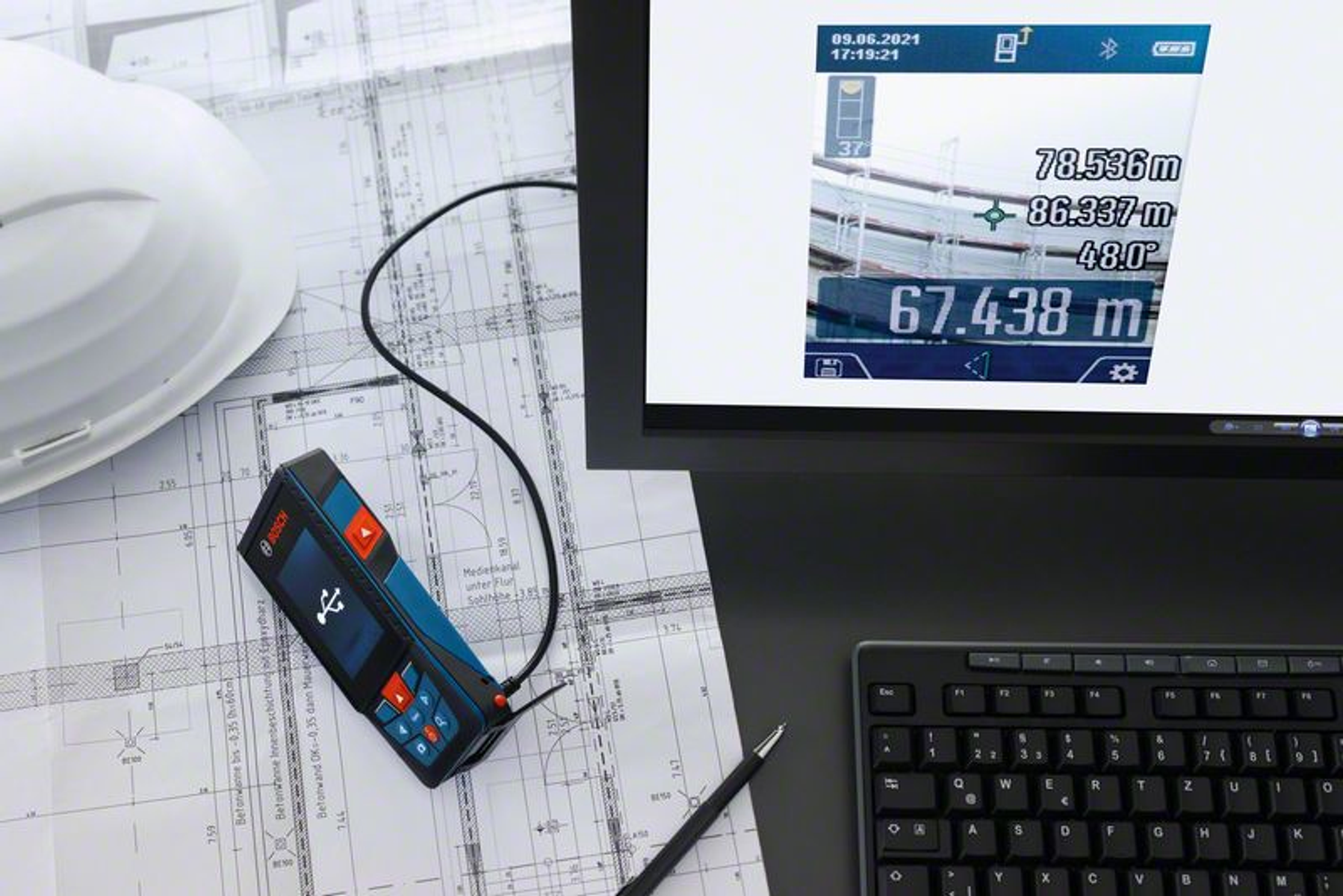 Un dispositivo di misurazione con display che mostra le misurazioni è appoggiato su progetti accanto a un computer e a una tastiera.