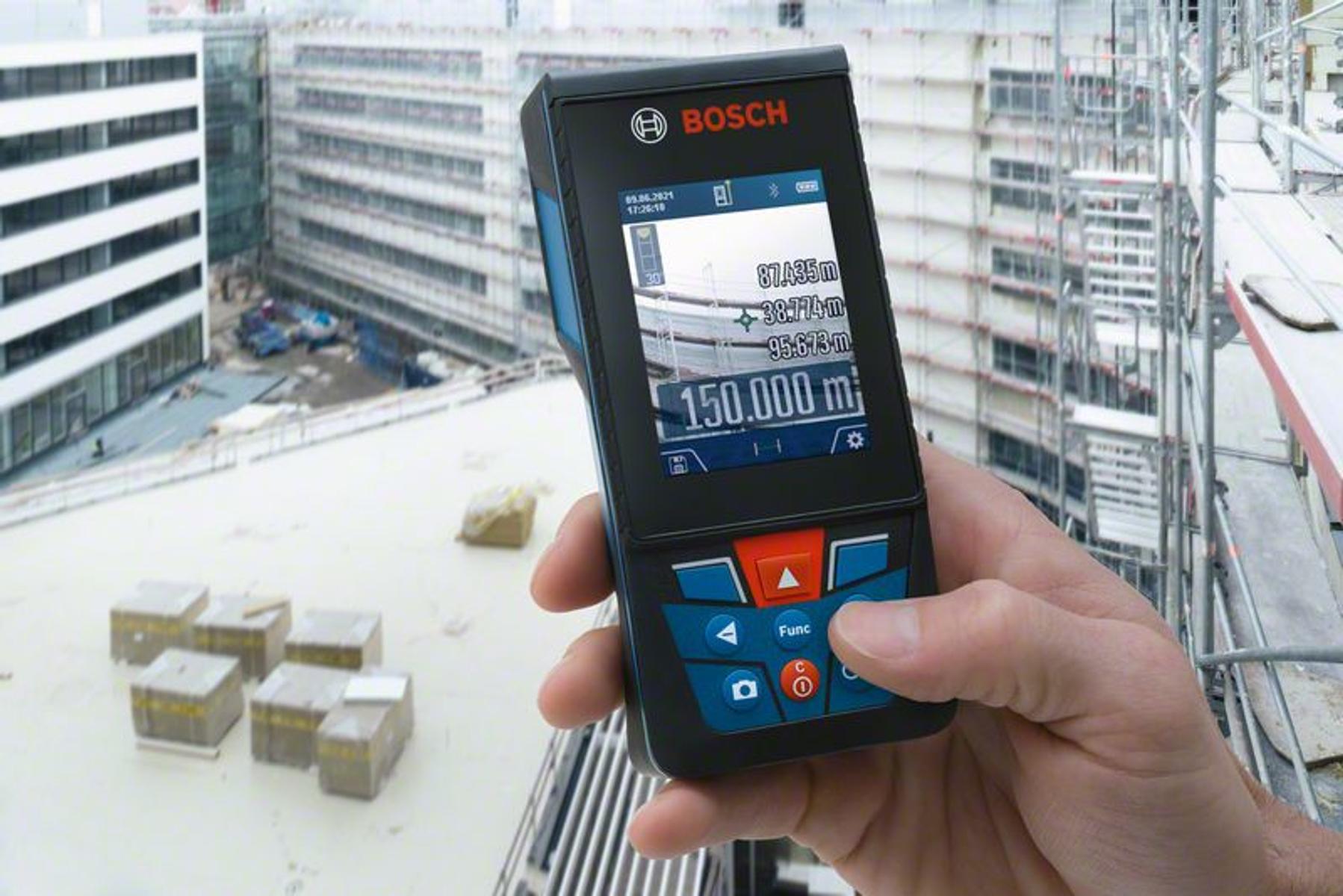 Una mano tiene in mano un telemetro laser Bosch. Edifici e ponteggi sullo sfondo. Scatole sul tetto.