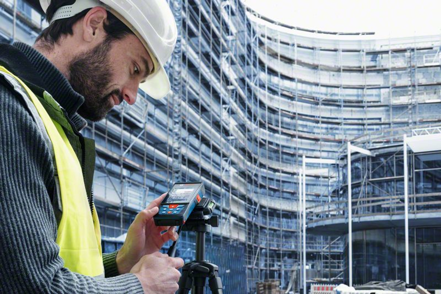 Un operaio edile con un casco bianco usa un dispositivo di misurazione. Un grande edificio curvo con ponteggi sullo sfondo.