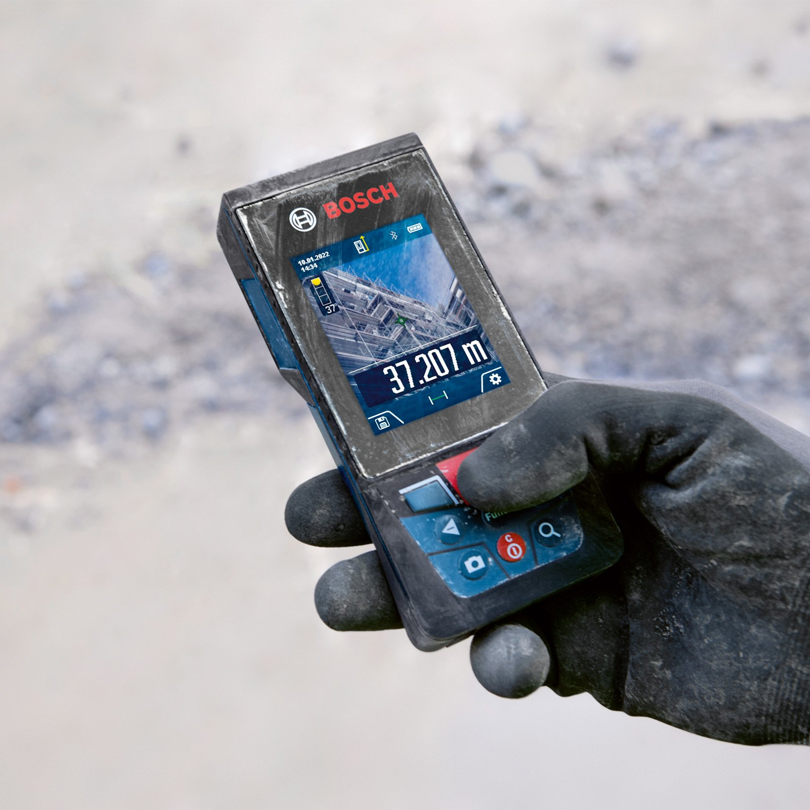 La mano tiene in mano un misuratore laser di distanza Bosch, che mostra 37,207 m. Lo sfondo è in cemento sfocato e detriti.