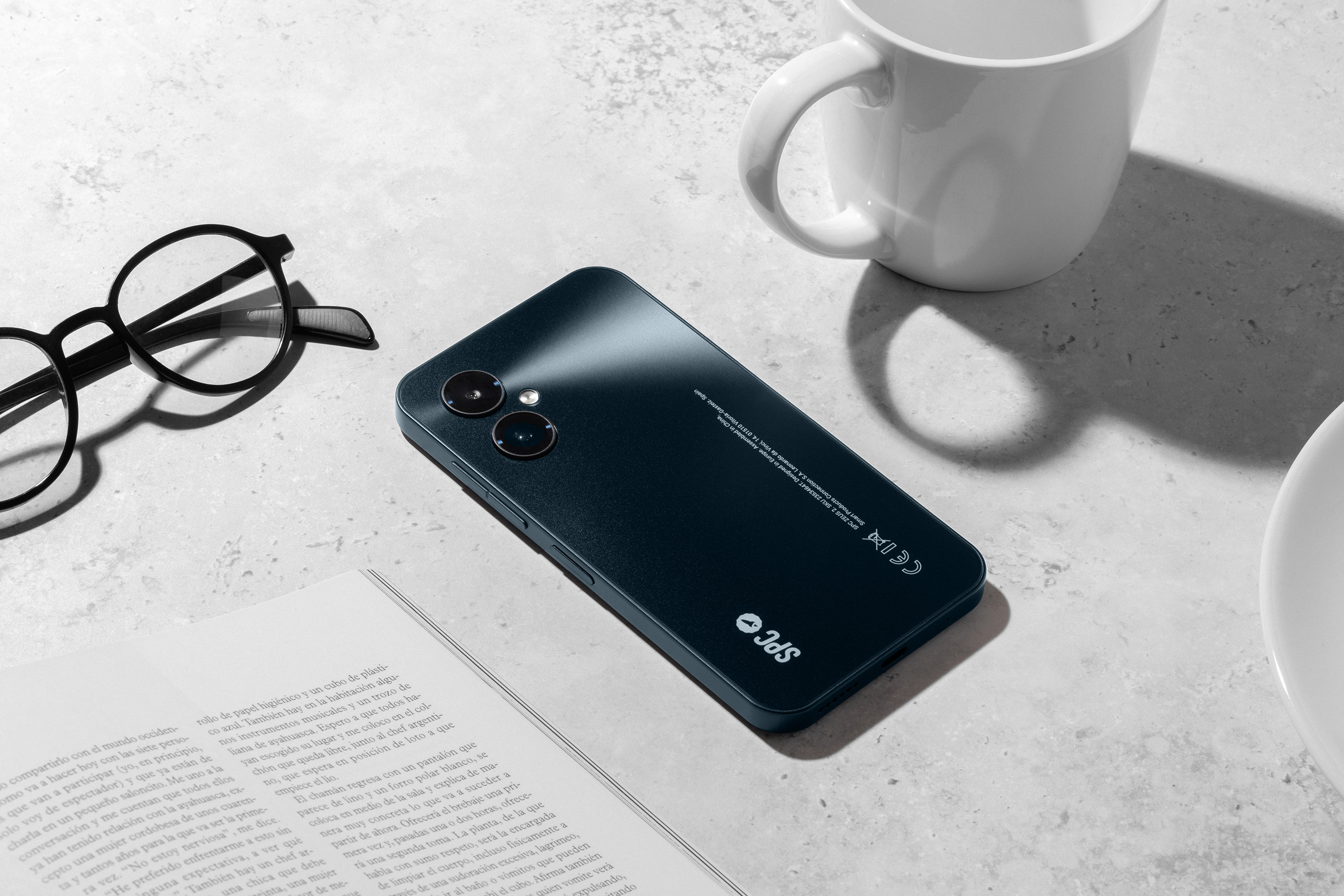 Ein schwarzes Smartphone, eine Brille, eine Tasse und ein offenes Buch liegen auf einer hellen Oberfläche.
