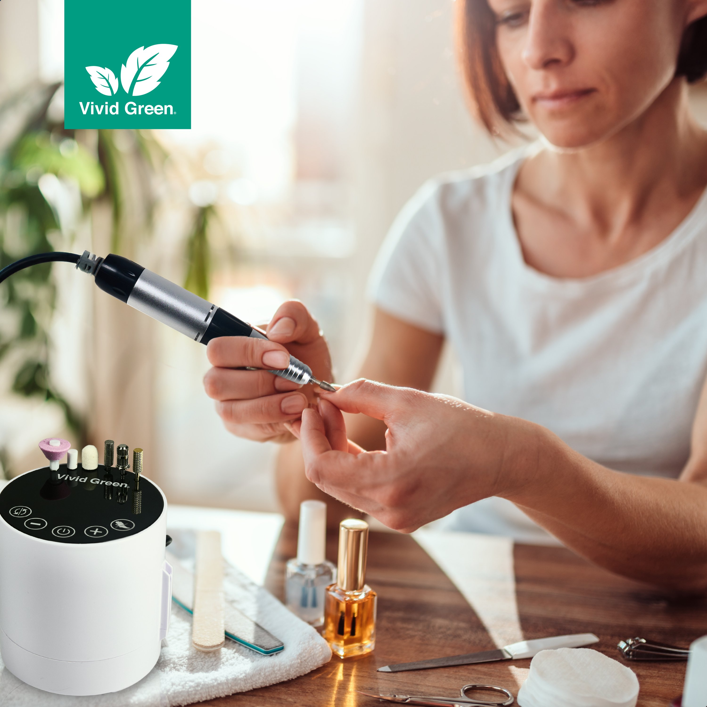 Woman using a nail drill. White machine and nail tools on the table. Vivid Green logo in the corner.