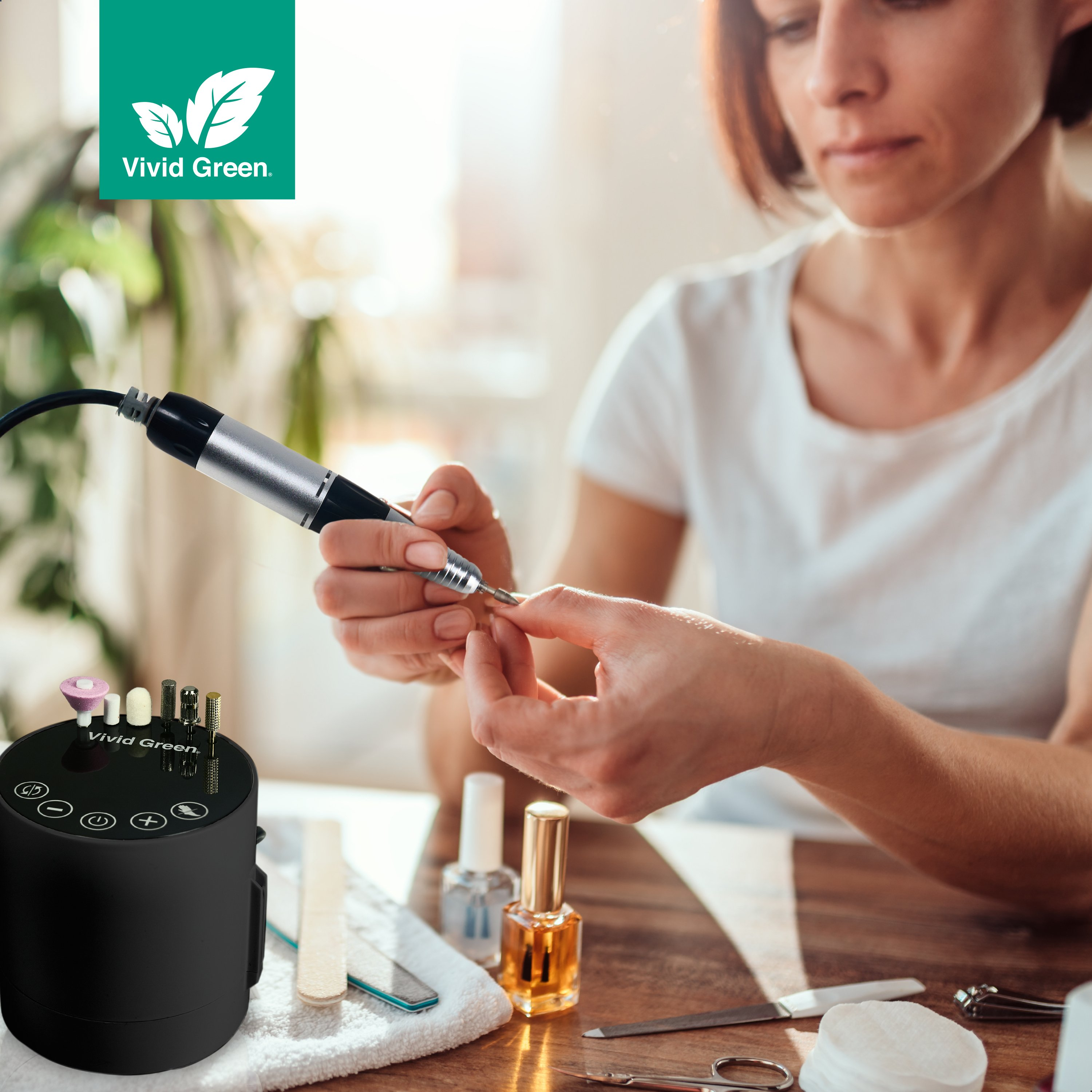 Woman using a nail drill. Nail tools and polish are on a table. A logo of Vivid Green is in the corner.
