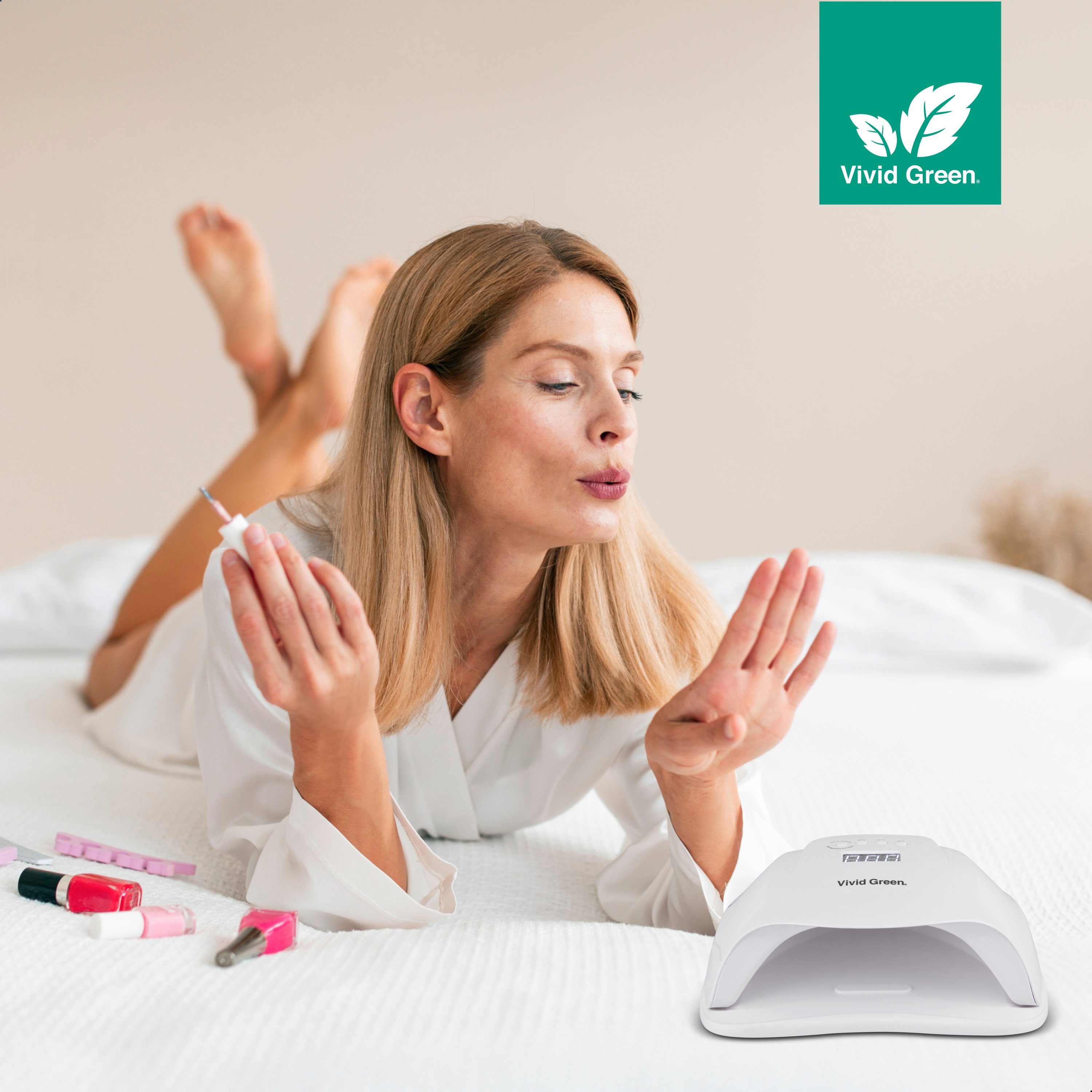 Woman on a bed is applying nail polish. Nail polishes and a UV lamp are visible on the bed. A green logo is in the corner.