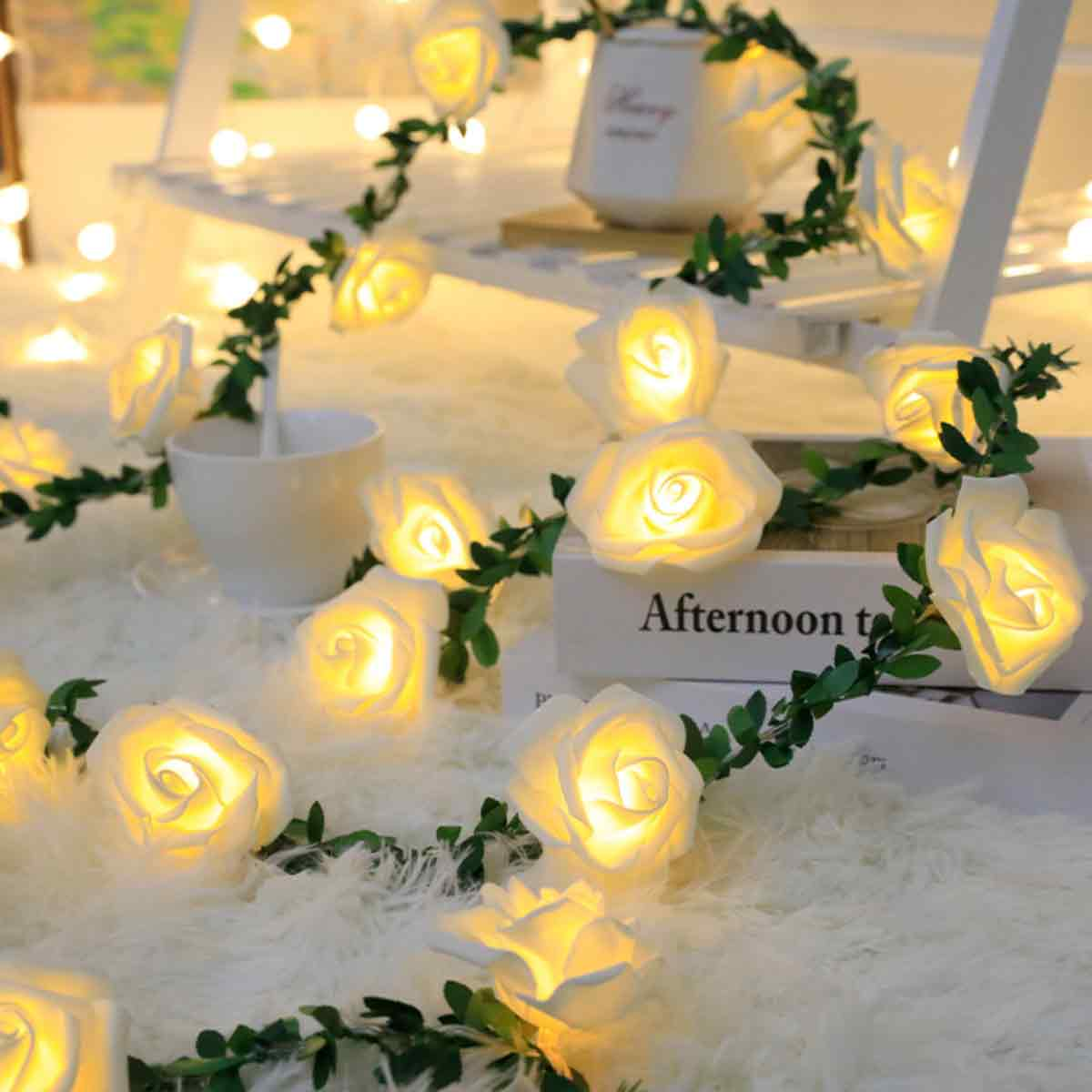 Weiße rosenförmige Lichter auf einer flauschigen weißen Oberfläche, mit grünen Ranken. Ein Buch mit "Afternoon t" und eine weiße Tasse sind sichtbar.