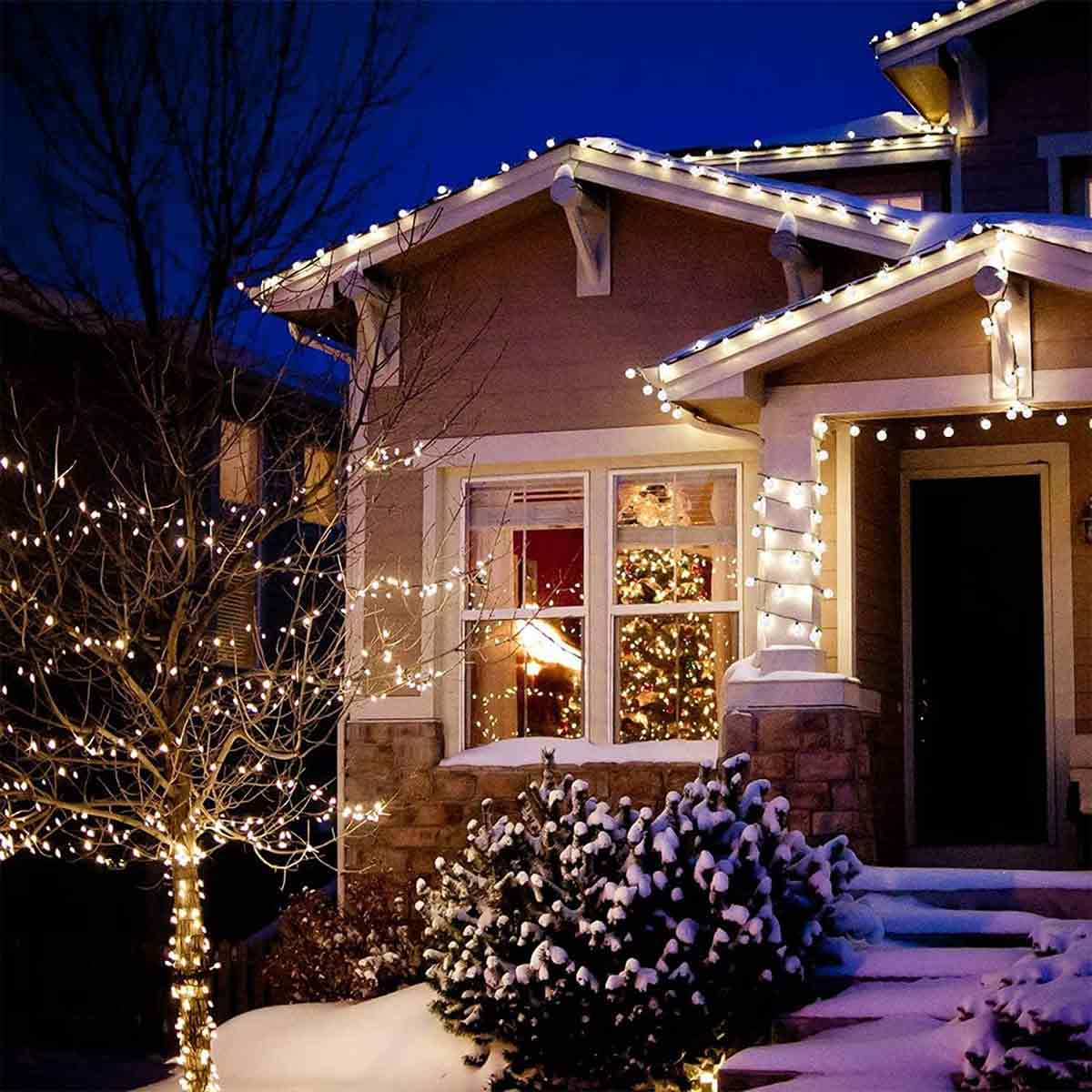 Ein Haus bei Nacht mit Weihnachtsbeleuchtung. Ein Baum und Büsche sind mit Lichtern und Schnee bedeckt. Das Haus hat einen Weihnachtsbaum im Fenster.