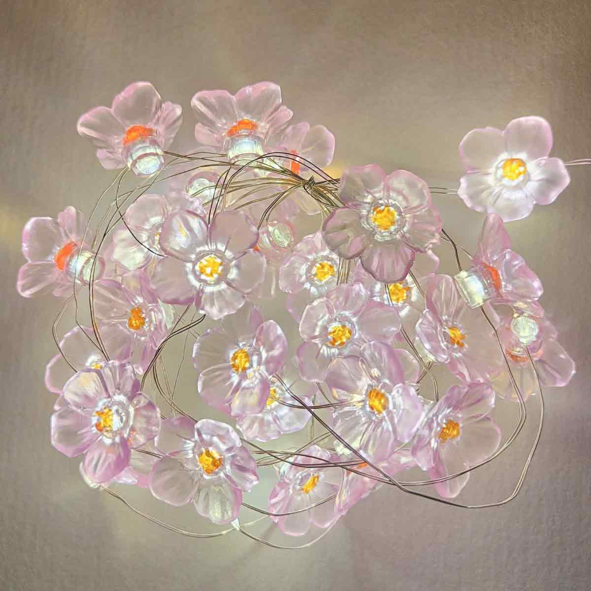 Nahaufnahme einer Lichterkette mit rosa Blüten. Jede Blüte hat eine gelbe Mitte. Drähte verbinden die Blumen. Der Hintergrund ist hellgrau.
