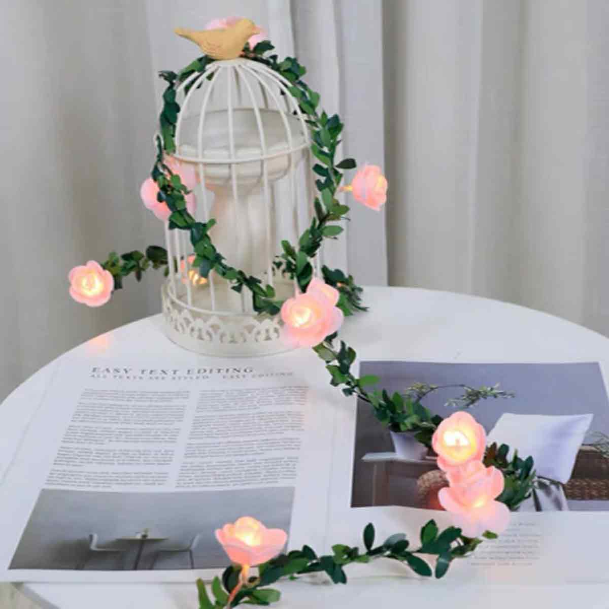 Ein weißer Vogelkäfig mit einem Blumengirlande aus rosa Rosen und grünen Blättern, geschmückt mit einer Vogelfigur, steht auf einem Tisch mit einer Zeitschrift.