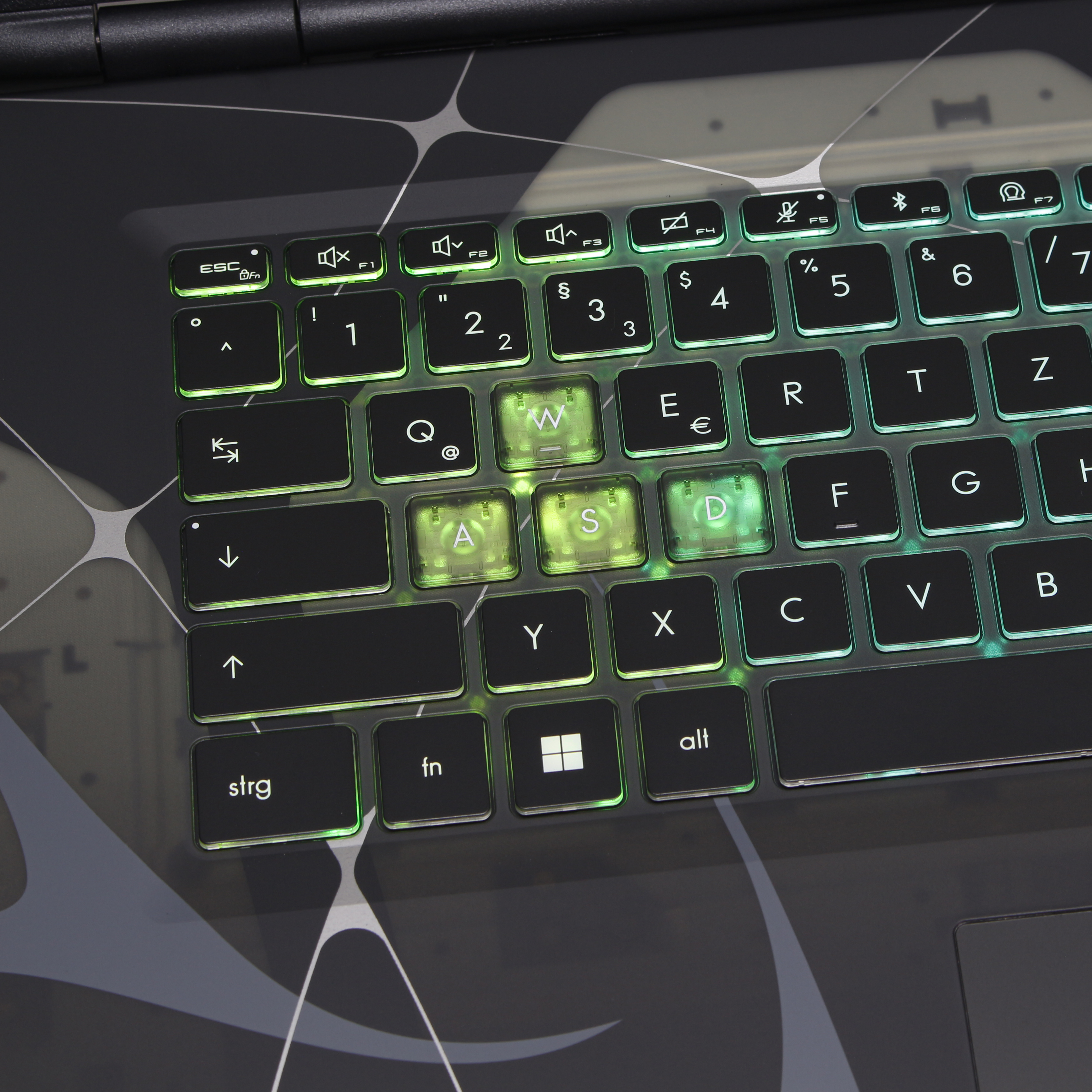 Nahaufnahme einer Laptop-Tastatur mit grün leuchtenden Tasten. Die Buchstaben WASD sind beleuchtet. Der Hintergrund hat graue Muster.