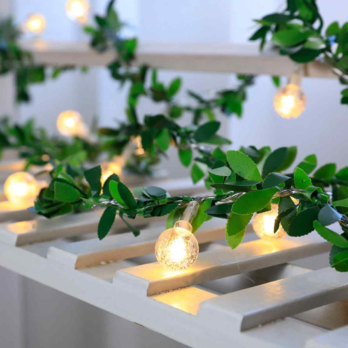 Eine Lichterkette mit klaren, runden Glühbirnen ist über weißen Holzlatten drapiert und mit grünen Blättern verflochten.