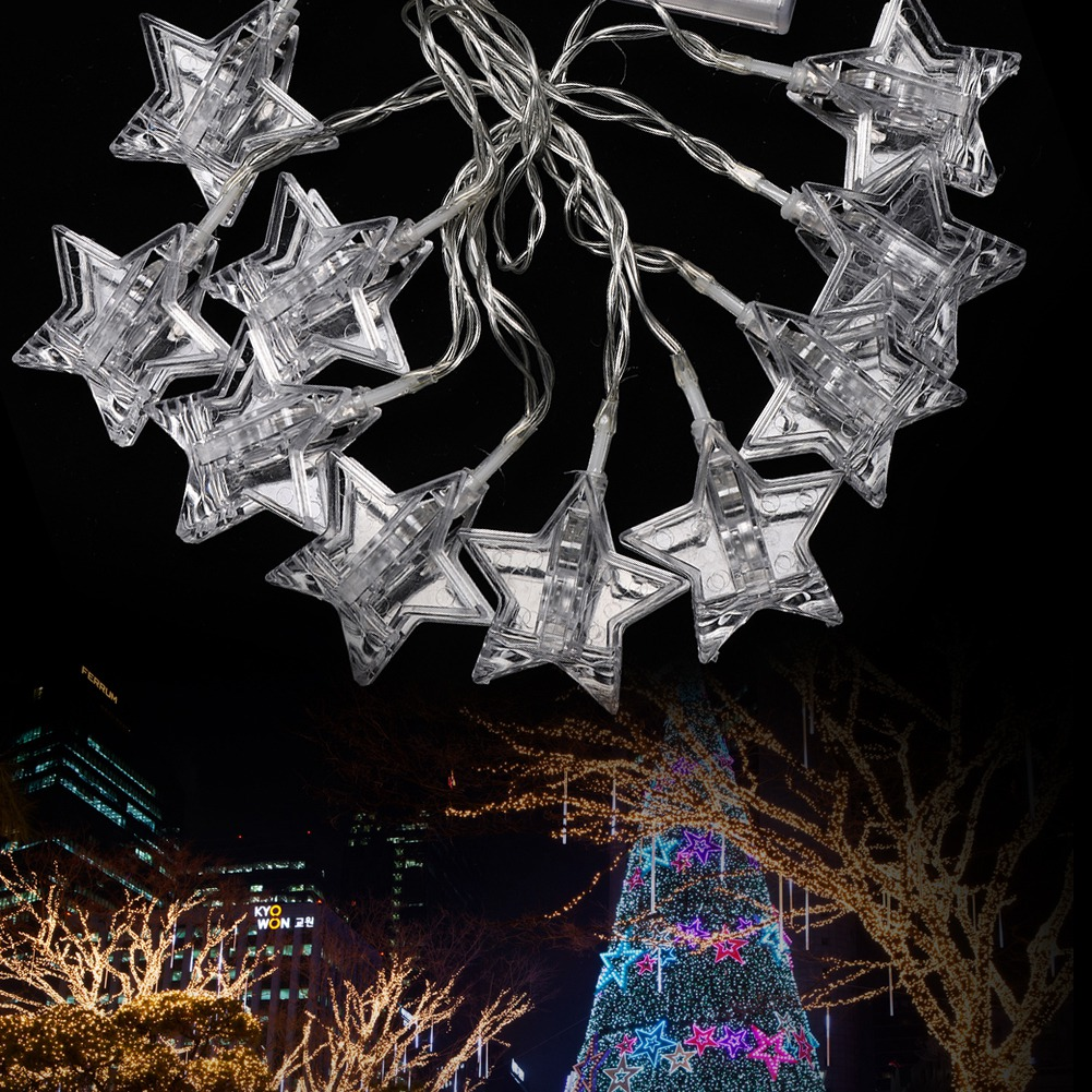 Lichterkette mit sternförmigen Glühbirnen. Der Hintergrund zeigt einen Weihnachtsbaum und Gebäude mit Lichtern bei Nacht.
