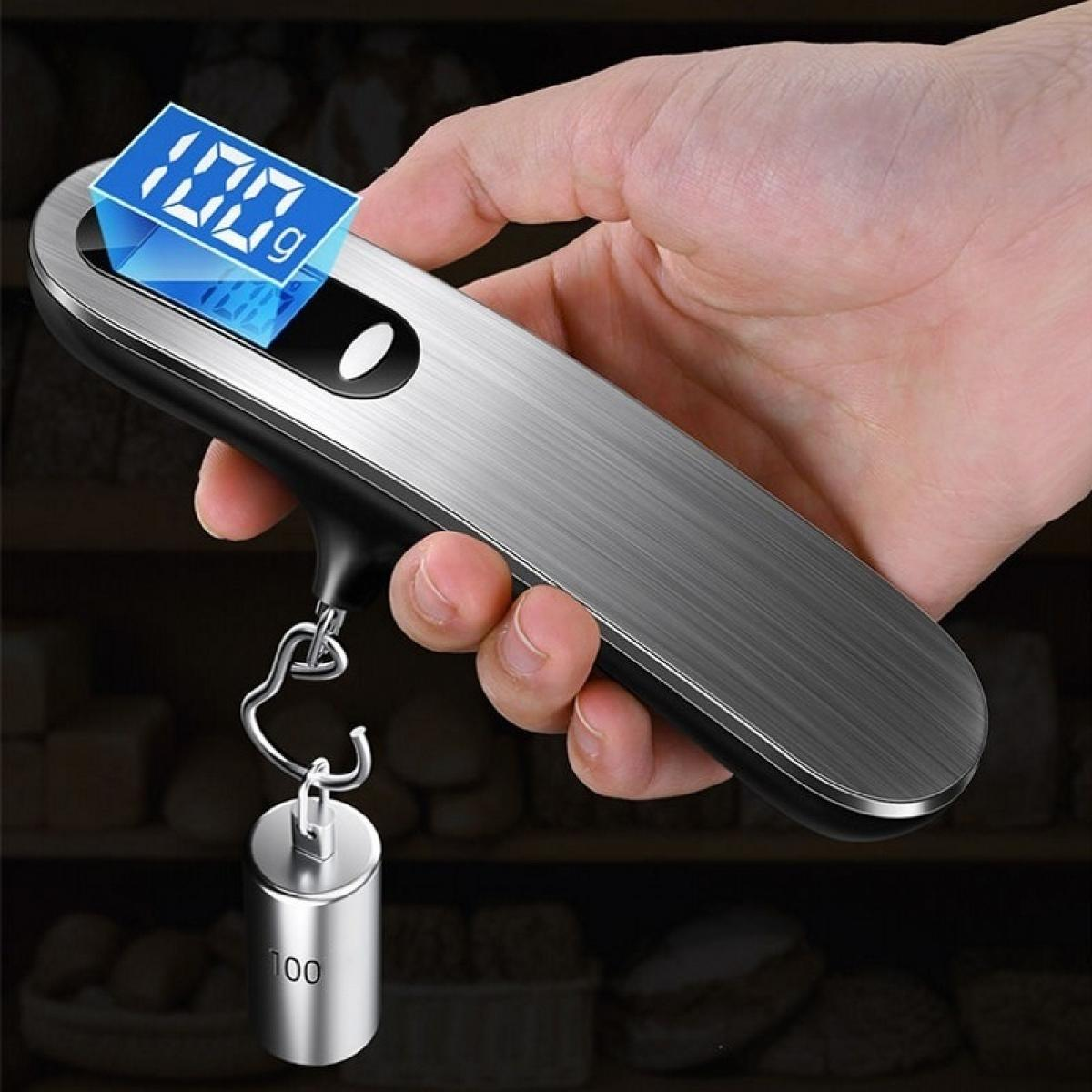 Eine Hand hält eine digitale Gepäckwaage mit 100g. Ein Metallgewicht hängt unten. Der Hintergrund zeigt ein verschwommenes Regal.