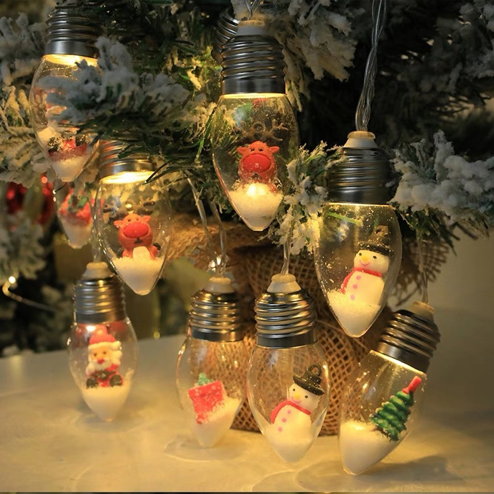 Weihnachtsbeleuchtung in Form von Glühbirnen, jede mit einer Figur: Schneemann, Weihnachtsmann, Rentier. Alle sind auf einem verschneiten Hintergrund.