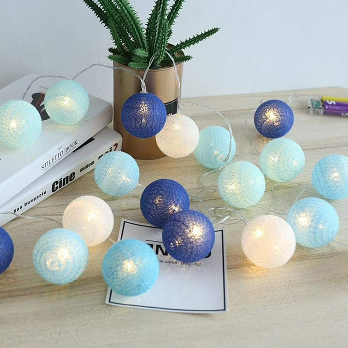 Lichterkette mit blau, weiß und hellblau leuchtenden Kugeln auf einem Holztisch, in der Nähe einer Pflanze und Büchern.