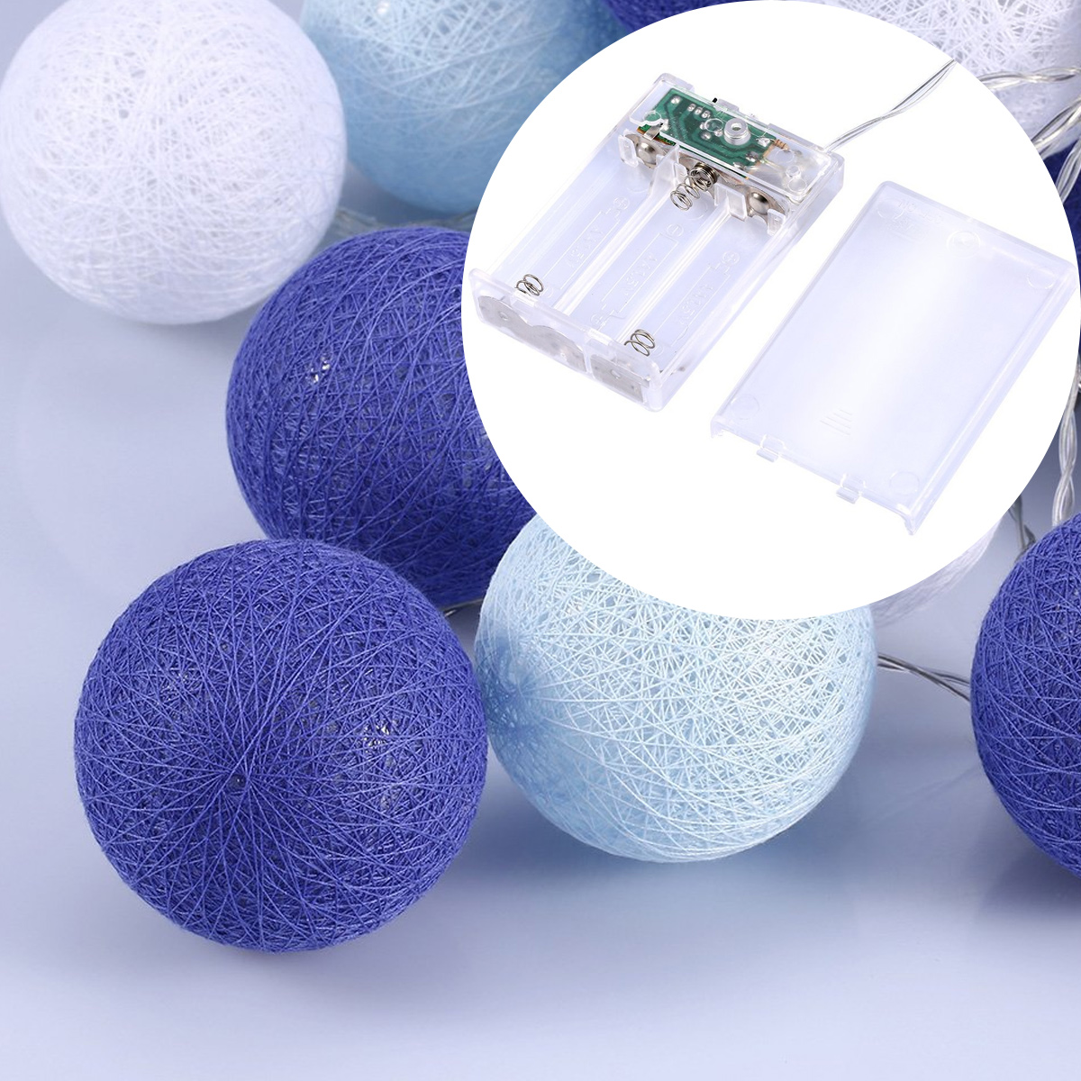 Nahaufnahme von Lichterketten mit blauen, weißen und violetten Garnkugeln. Eine transparente Batteriepackung ist abgebildet. Der Hintergrund ist hellblau.