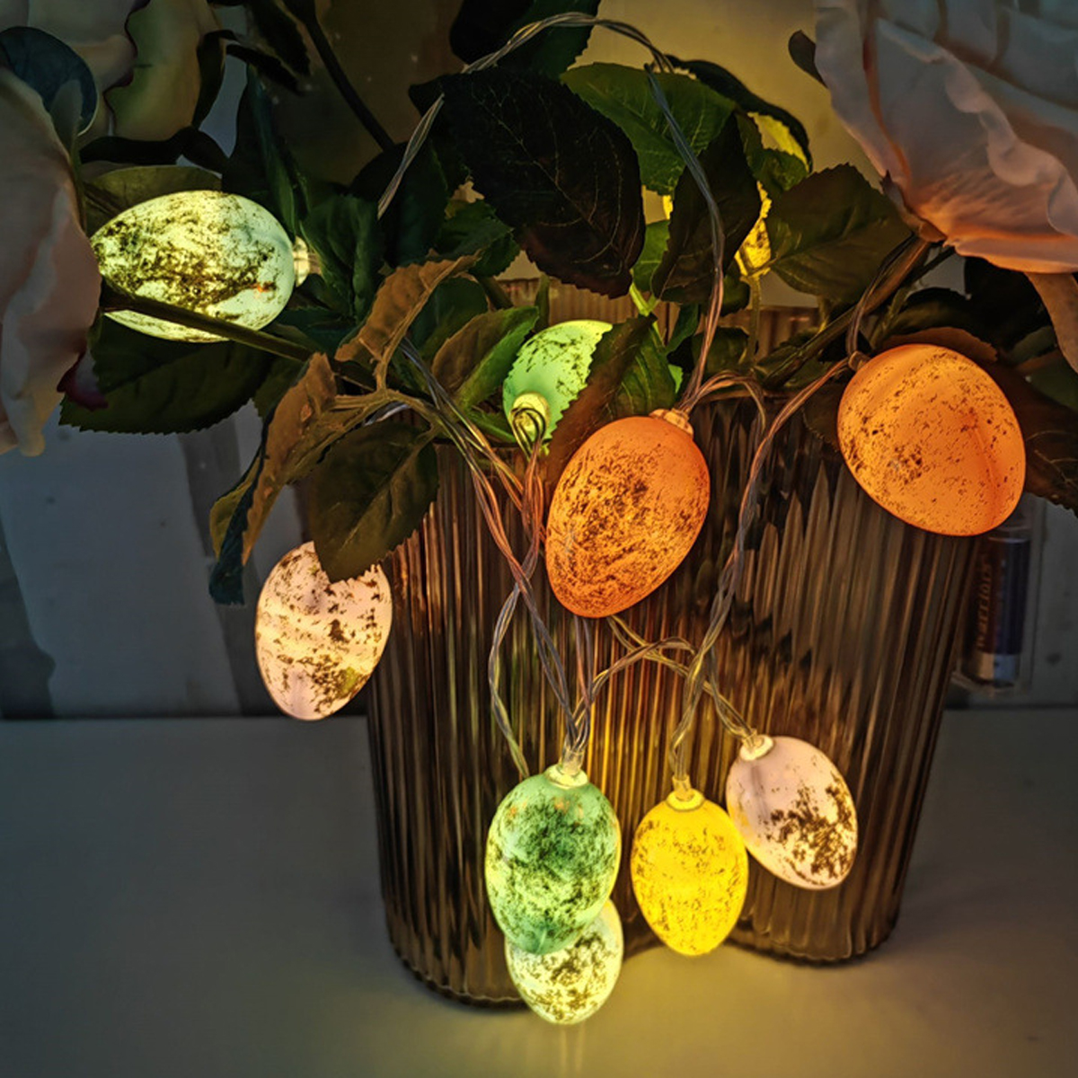 Ostereierlichter in einer Vase mit grünen Blättern und einer weißen Rose. Eier sind in verschiedenen Farben: orange, grün, gelb und weiß.