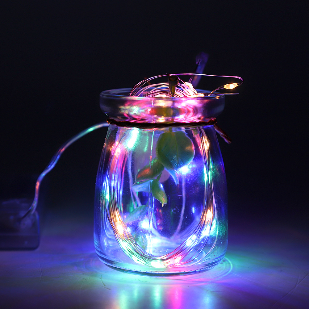 Ein Glas mit Feenlichtern darin. Grüne Blätter schauen heraus. Die Lichter leuchten in verschiedenen Farben vor dunklem Hintergrund.