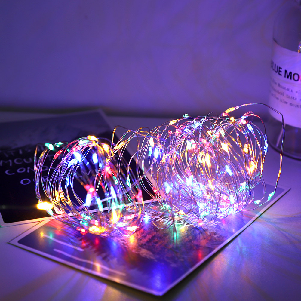 Nahaufnahme von bunten Lichterketten, die auf einer reflektierenden Oberfläche leuchten. Im Hintergrund eine verschwommene Glasflasche. Die Szene ist in violettes Licht getaucht.