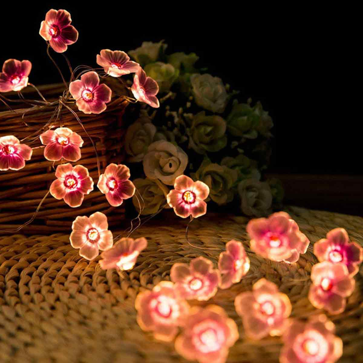 Beleuchtete Blumengirlande. Rosa Blüten mit weißen Zentren liegen auf einer gewebten Oberfläche, im Hintergrund ein Blumenstrauß.