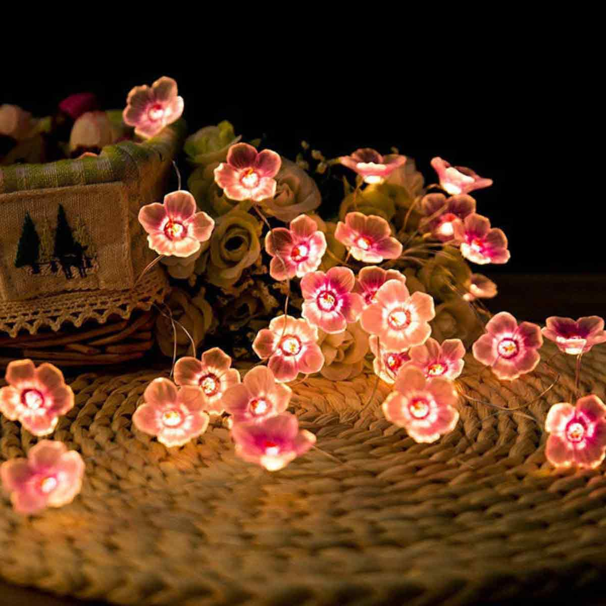 Nahaufnahme von Blumenschnur-Lichtern. Rosa Blumen liegen auf einer gewebten Oberfläche. Einige Blumen sind im Korb mit weißen Blumen.