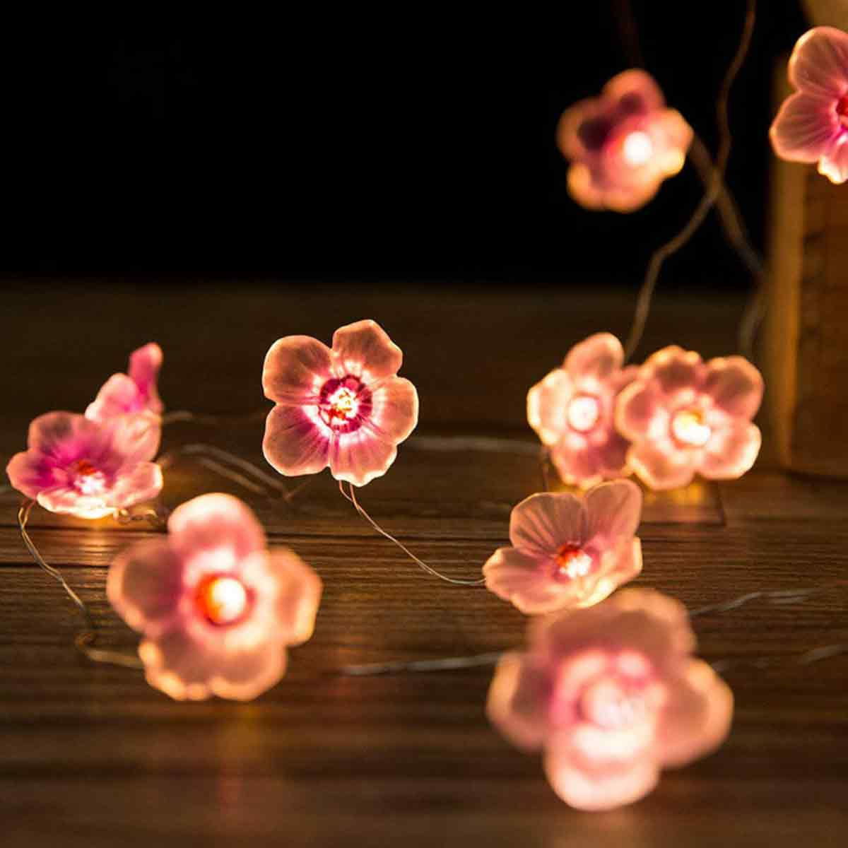 Nahaufnahme von blütenförmigen Lichterketten. Rosafarbene Blüten mit leuchtenden Zentren liegen auf einer Holzoberfläche vor dunklem Hintergrund.