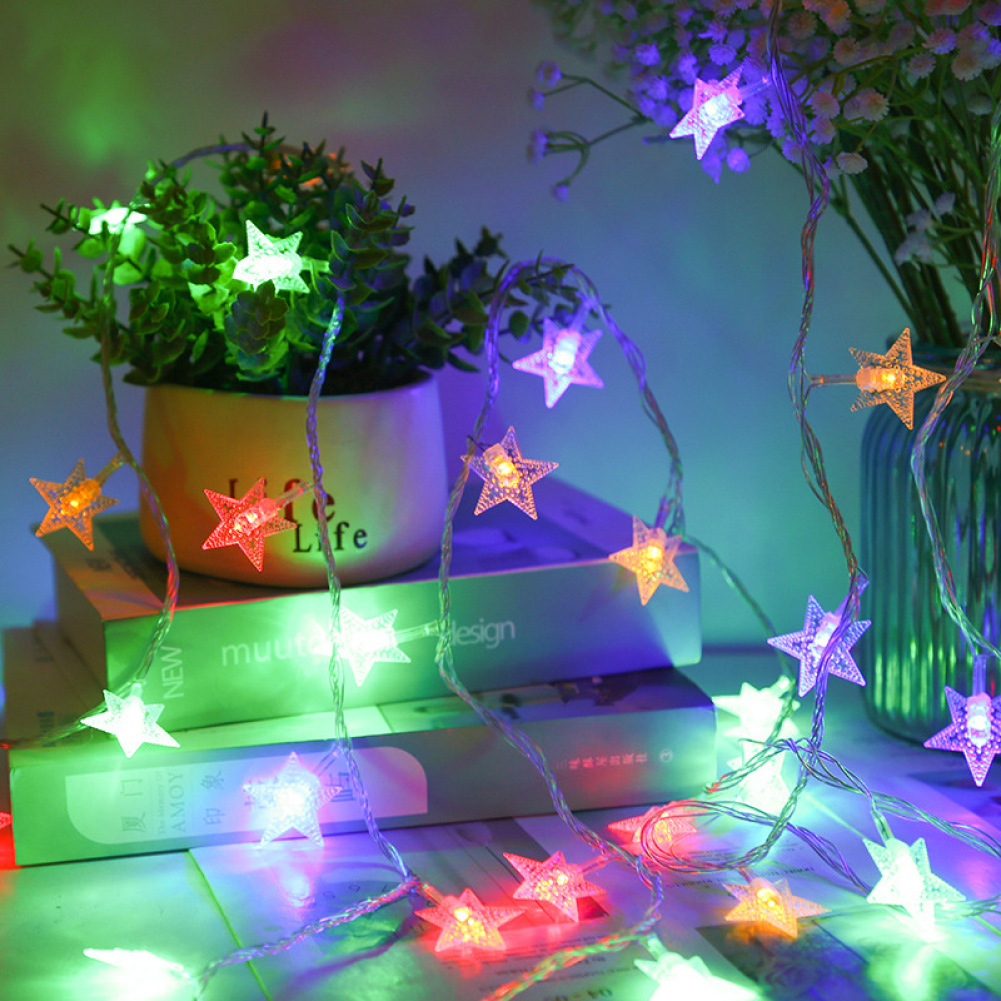 Eine Lichterkette in Sternform liegt über Büchern, Pflanzen und einer Vase. Die Sterne leuchten in verschiedenen Farben und schaffen Feststimmung.