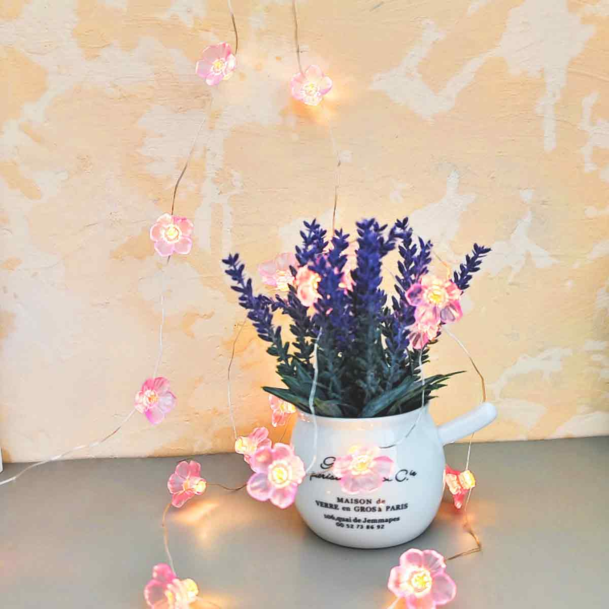 Ein weißer Topf mit Lavendel und rosa Blumenlichtern. Die Lichter sind um den Lavendel herum angebracht. Im Hintergrund ist eine hellgelbe Wand.