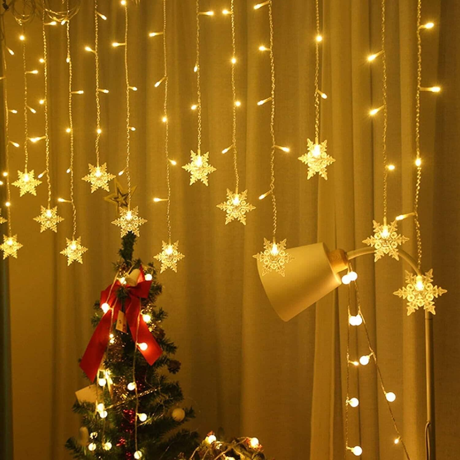 Ein geschmückter Weihnachtsbaum mit roter Schleife, beleuchtet von Lichterketten und Schneeflockenlichtern. Eine Lampe und ein Vorhang im Hintergrund.