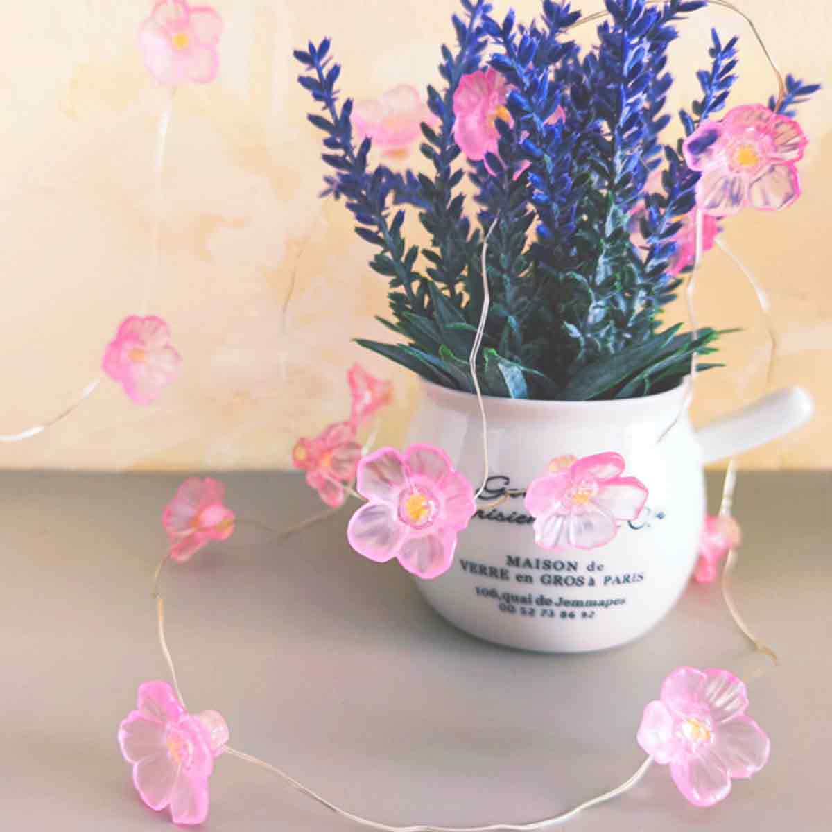 Ein weißer Topf mit Lavendel und pinken Blumenlichtern. Der Topf hat Text darauf. Der Hintergrund ist eine Wand.
