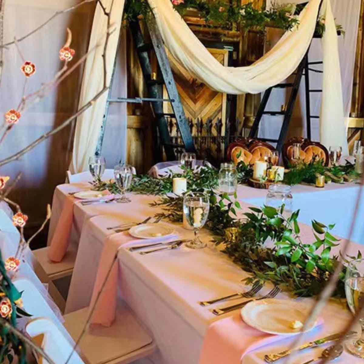Dekorierter Hochzeitstisch mit rosa Tischdecke, Gläsern, Tellern und Grünpflanzen. Hintergrund umfasst eine Leiter und Blumengestecke.