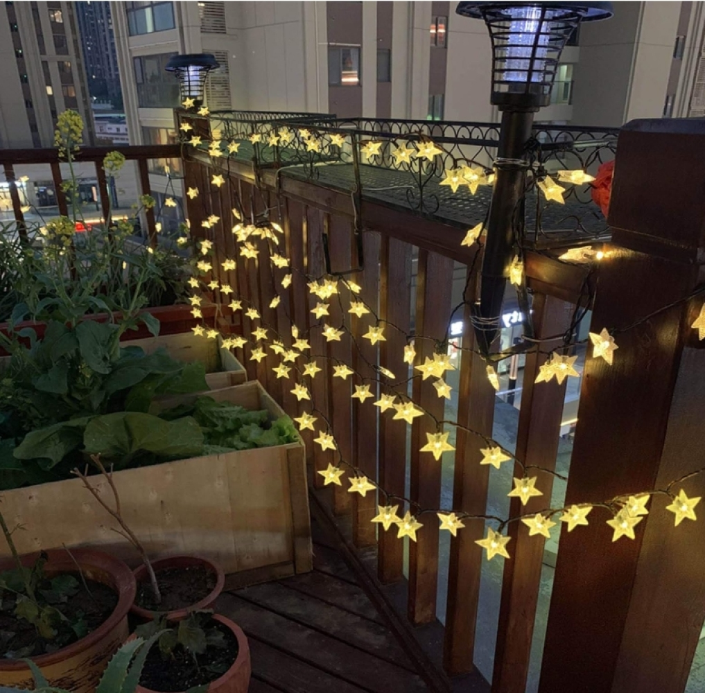 Ein Balkon bei Nacht mit sternförmigen Lichtern. Holzpflanzkästen mit Pflanzen im Vordergrund, Gebäude im Hintergrund.