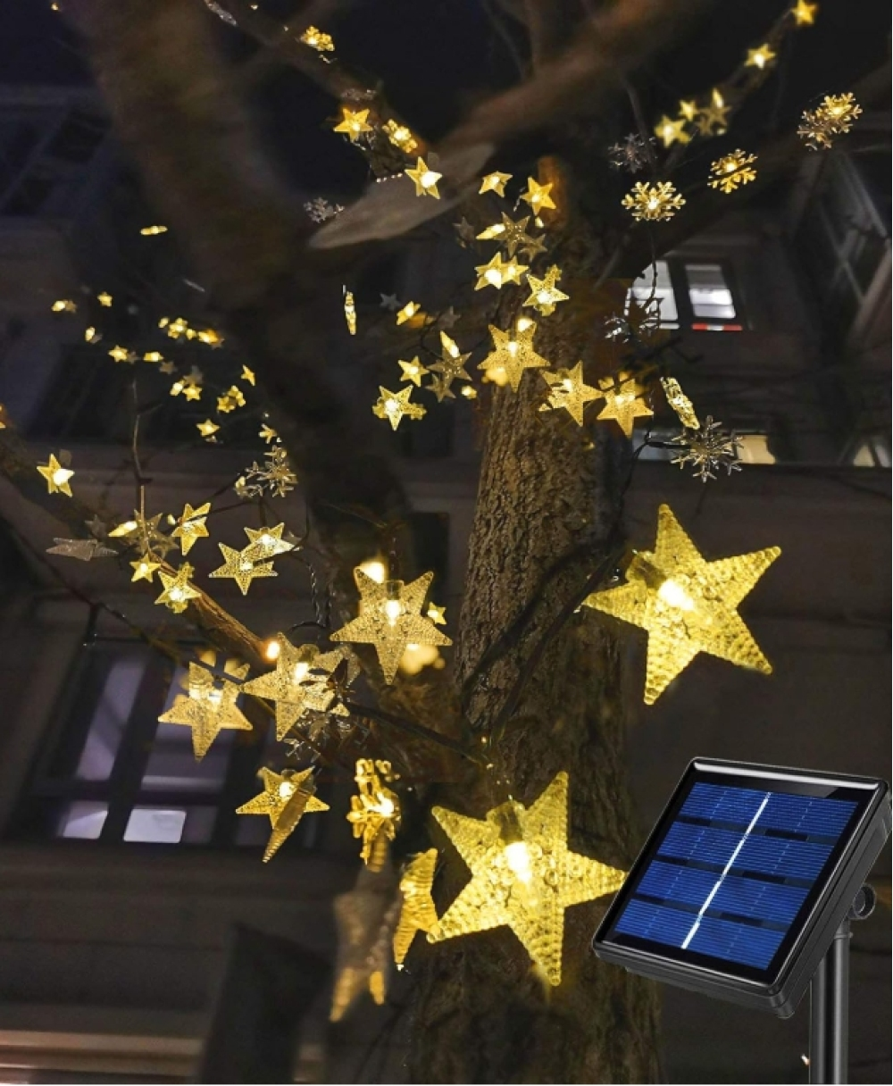 Ein Baum mit sternförmigen Lichtern geschmückt. Im Vordergrund befindet sich ein Solarpanel. Nachtszene mit Gebäude im Hintergrund.