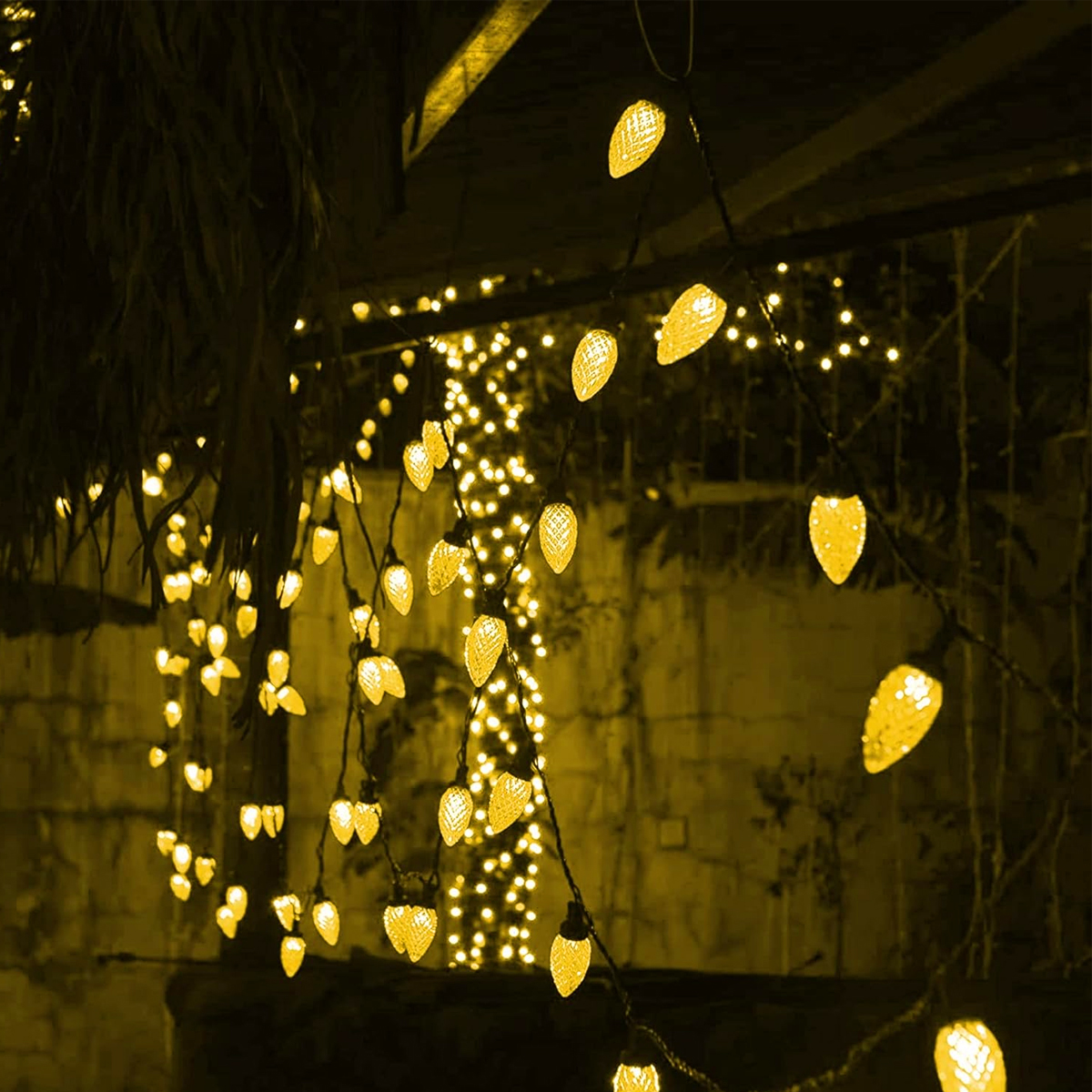 Lichterketten mit gelben Glühbirnen hängen von einem dunklen Dach. Im Hintergrund befinden sich weitere gelbe Lichter. Dunkle, blattreiche Pflanze links oben.