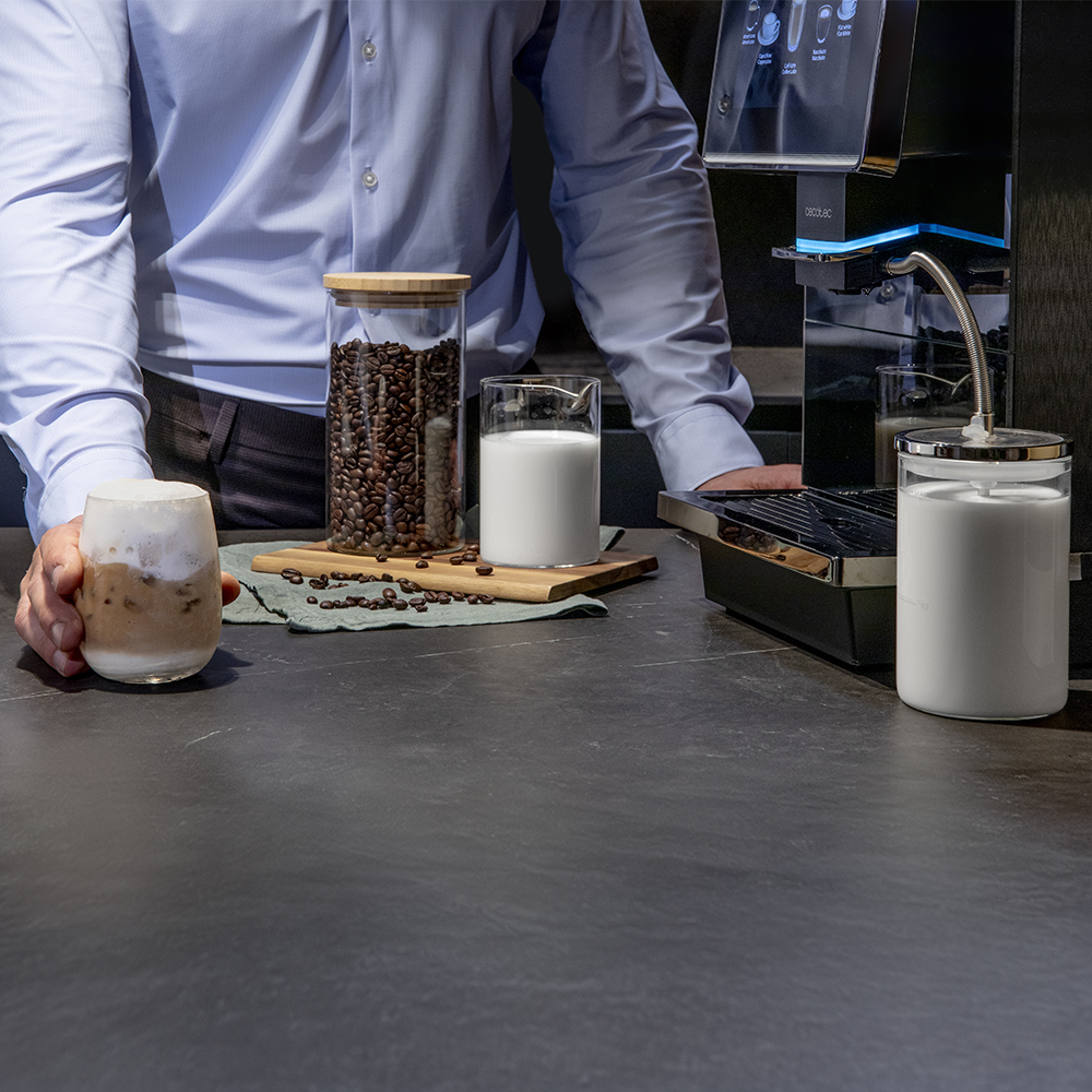 Una persona está detrás de un mostrador con granos de café, leche y una cafetera. Un vaso de café helado en primer plano.