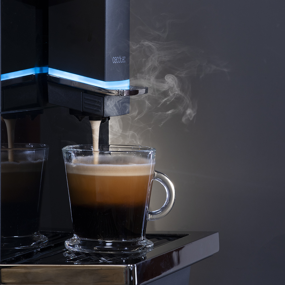 Cafetera vertiendo café en una taza de vidrio. El vapor se eleva. El café tiene capas oscuras y claras. Otra taza a la izquierda.