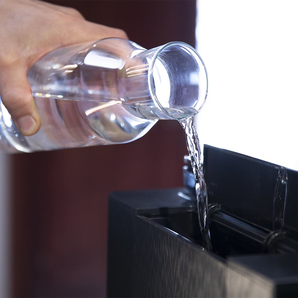 Una mano vierte agua de una jarra de vidrio transparente en un recipiente negro. El chorro de agua es claro, con un fondo rojo.