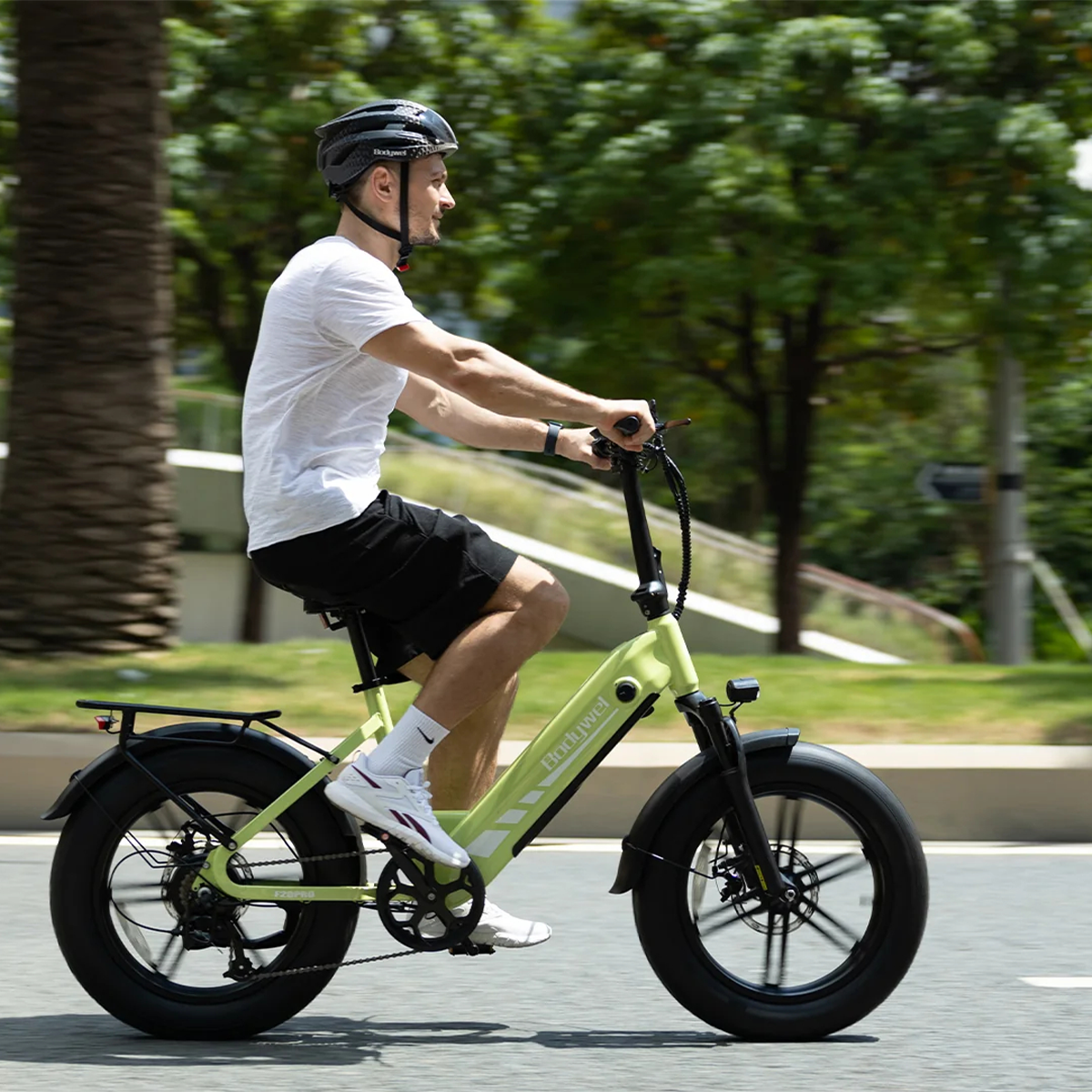 Mężczyzna jedzie jasnozielonym rowerem elektrycznym po drodze. Ma na sobie kask, białą koszulkę i szorty. W tle drzewa.