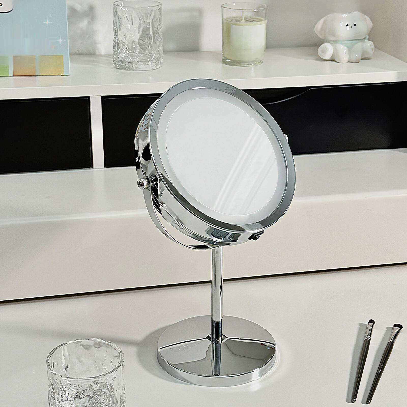 Ein Schminkspiegel steht auf einer weißen Oberfläche mit Make-up-Pinseln. Im Hintergrund sind Glas, eine Kerze und eine weiße Hundefigur zu sehen.