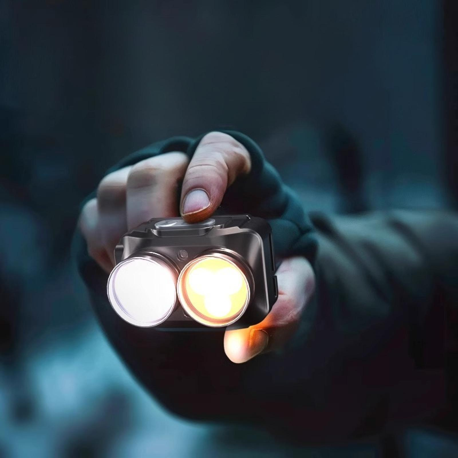 Eine Hand, die eine kleine schwarze Taschenlampe mit zwei hellen Lichtern hält, eines weiß und eines gelb, in dunkler Umgebung.