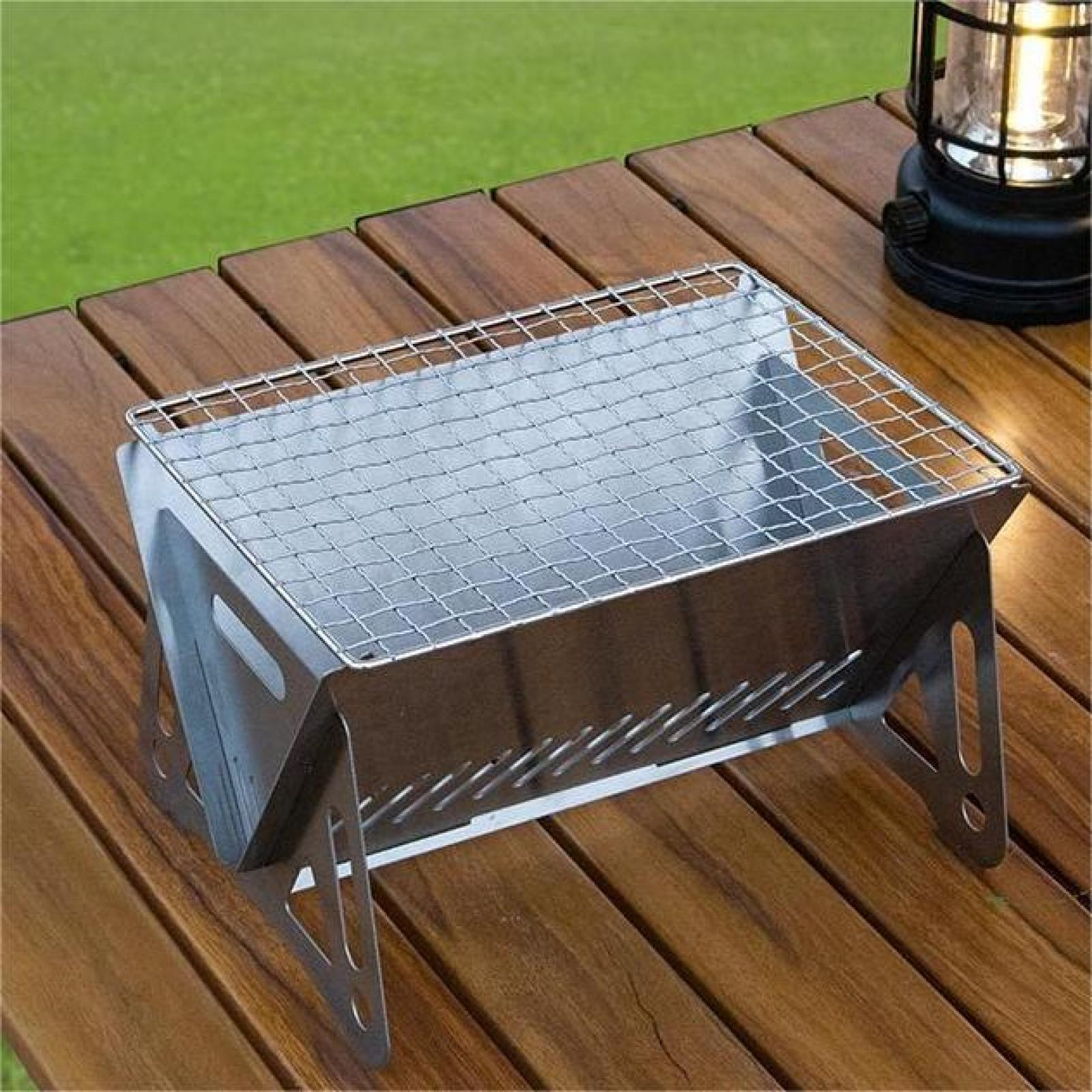 Ein tragbarer Grill aus Edelstahl steht auf einem Holztisch, im Hintergrund eine beleuchtete Laterne und grünes Gras.
