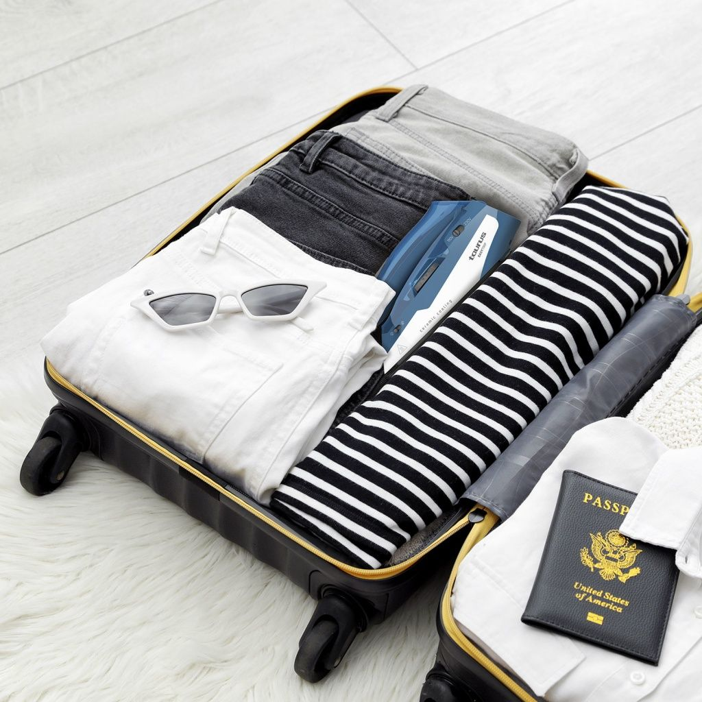 Maleta abierta con ropa, gafas de sol y pasaporte. Incluye jeans, camisa a rayas y plancha azul. Lista para viajar.